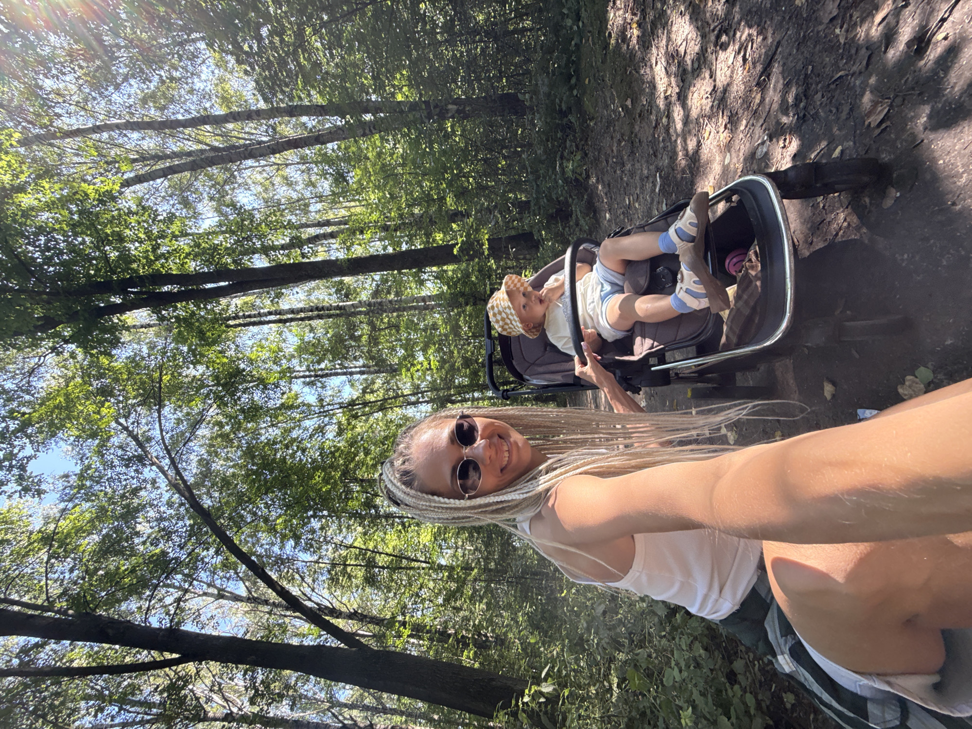 A young woman with blonde braided hair and sunglasses takes a selfie in a sun-dappled forest path, holding her phone with her left hand. She is wearing a white tank top and a plaid skirt. Beside her, a child in a checkered hat sits in a stroller, looking at their hand. The stroller has a dark frame and brown fabric. The background is filled with tall trees and lush green foliage, suggesting a natural, wooded environment. The overall impression is a casual outing in a park or forest.