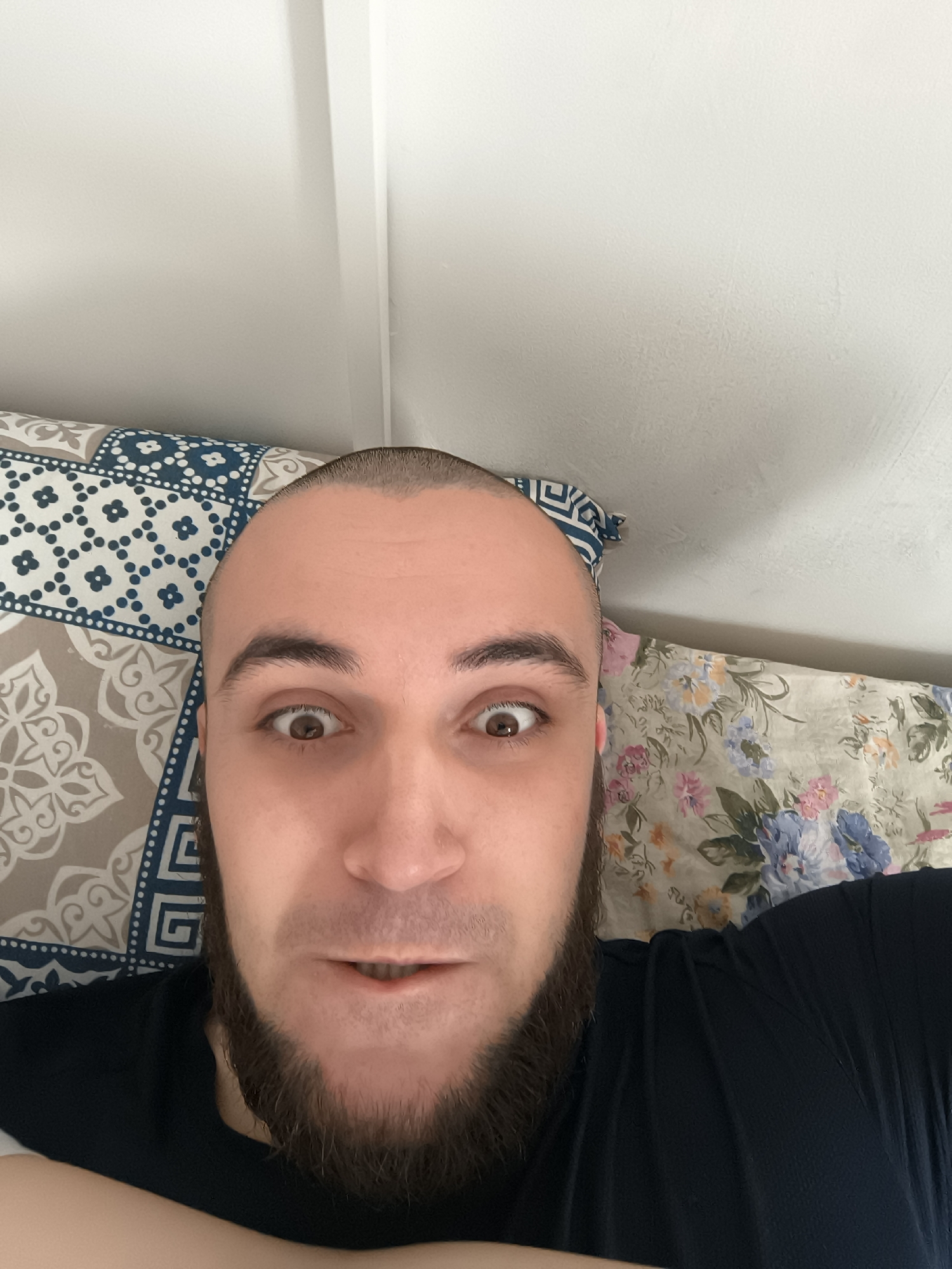 A bald, fair-skinned man with a dark beard is lying down, taking a selfie from a low angle. His eyes are wide open and he appears to be looking directly at the camera. He is wearing a black shirt. He is resting on two pillows; one with a blue and beige geometric pattern, and the other with a floral pattern. The background shows a white wall and a white door frame. There is no information available about the location being Veliko Tarnovo, Bulgaria.