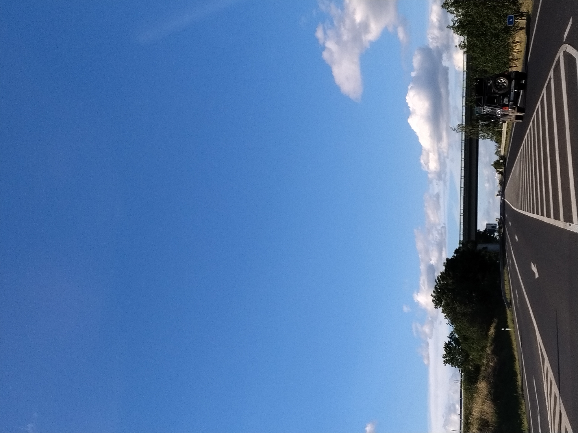 The image depicts a highway scene under a clear blue sky with scattered white clouds. In the foreground, a dark-colored off-road vehicle with a spare tire mounted on the back is driving on the right lane of the asphalt road. The road has clear white lane markings, including directional arrows. In the background, there is a highway overpass and a line of trees and vegetation on both sides of the road. A blue sign with the number "4" is visible on the right side of the road. The scene appears to be in a rural or suburban setting. The location is identified as Předotice, Czechia. There are no people visible in the image.