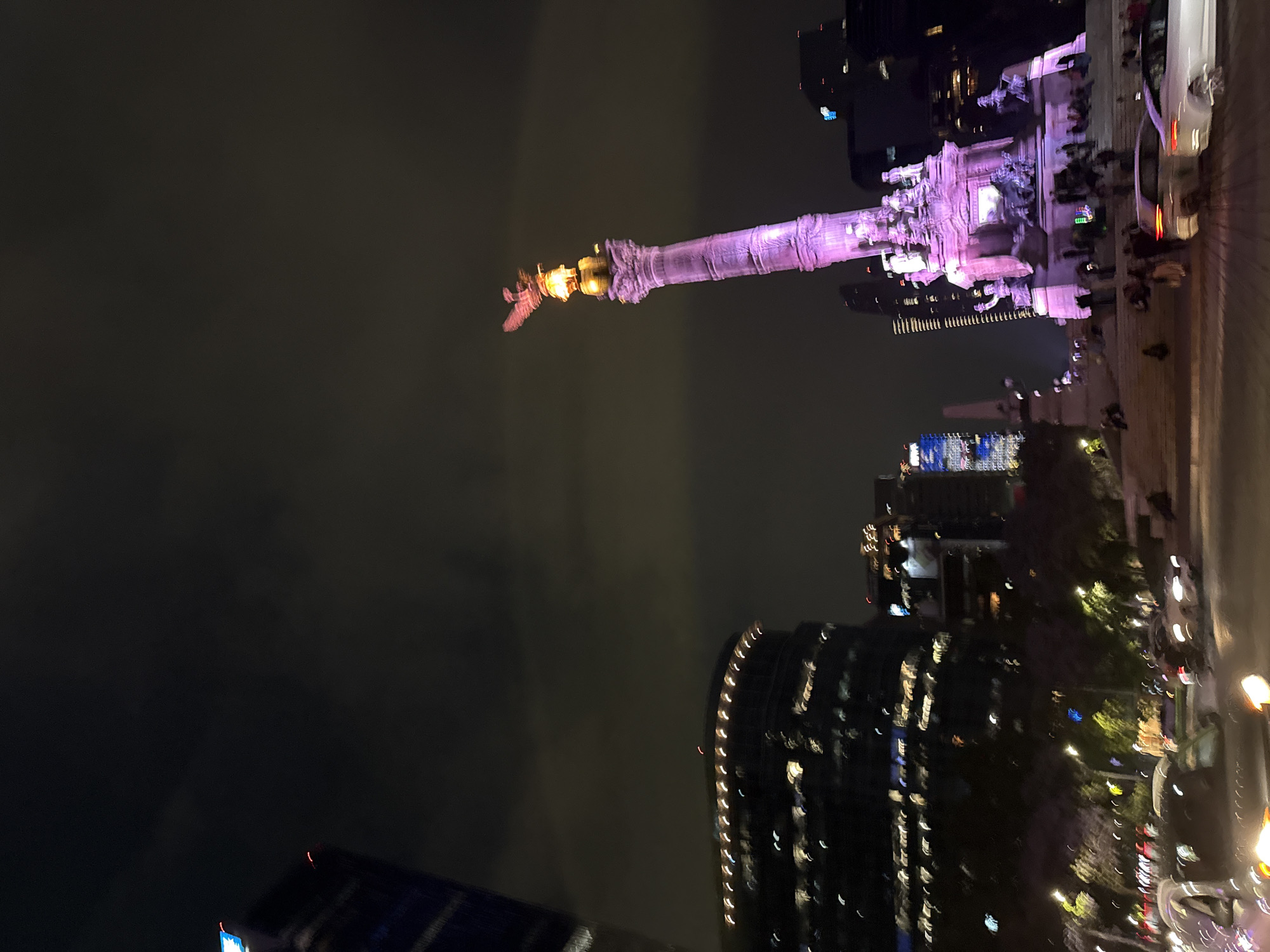 At night, the Angel of Independence monument glows brightly in the city of Mexico City, Mexico. The monument is a towering column topped with a golden angel, bathed in pink and purple light. Below the angel is a platform with statues and carvings. On the right, a sidewalk runs alongside the monument, with a crowd of people gathered around. Cars line the street in front of the sidewalk, with their headlights adding to the scene's glow. Buildings with lit windows flank the monument. Dark clouds dominate the sky.