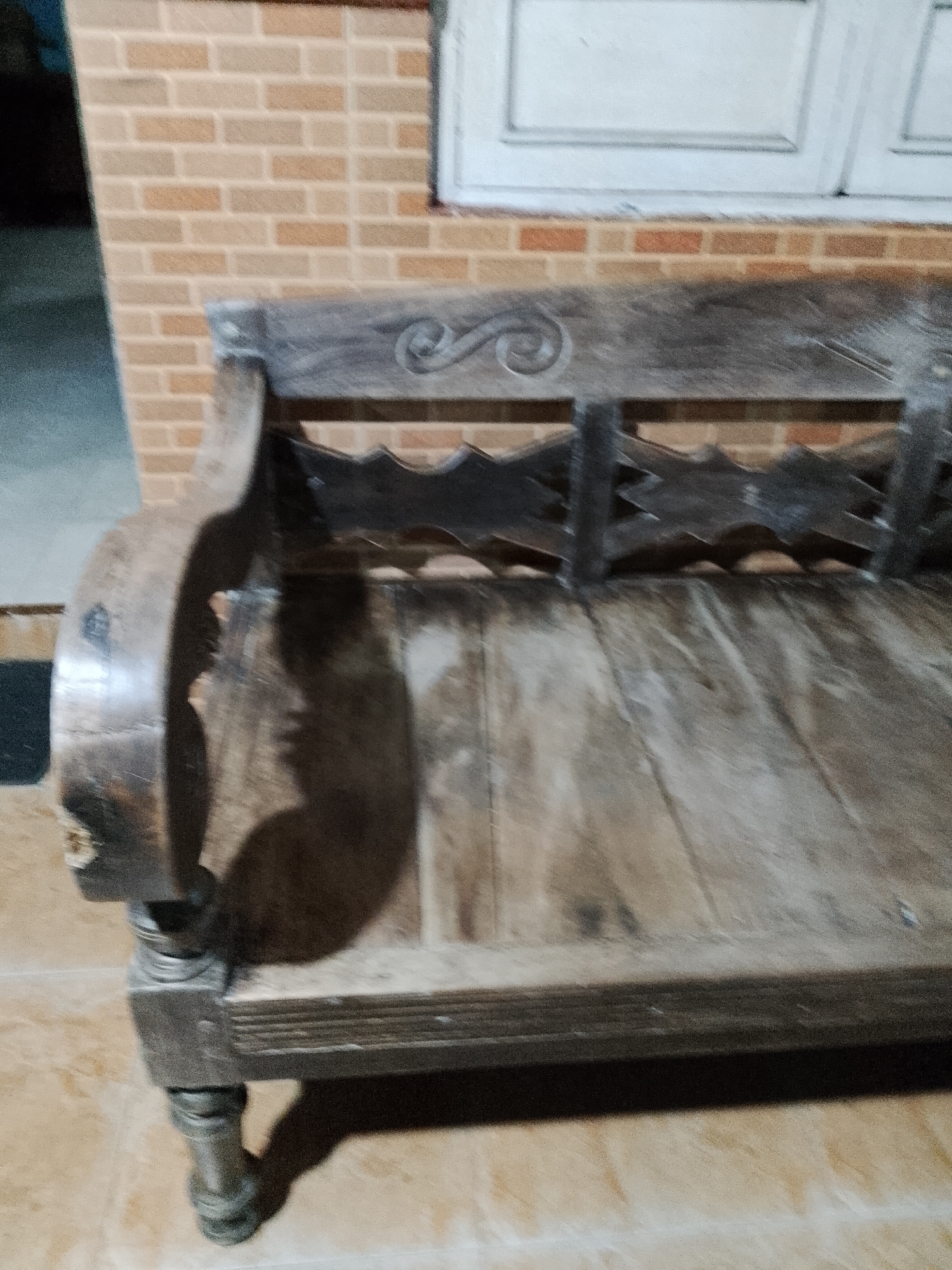 The image shows a close-up, slightly blurry view of an old, weathered wooden bench in an outdoor setting. The bench has a carved backrest with a swirling pattern and a seat made of several wooden planks. The bench's legs are intricately carved and appear to be made of dark wood, possibly metal. The background consists of a brick wall and a portion of a window with white panes. The ground beneath the bench is tiled. The lighting suggests it is daytime, with a shadow cast by the bench on the ground. The scene is likely in Kecamatan Pati, Indonesia, given the context provided. There are no people visible in the image.