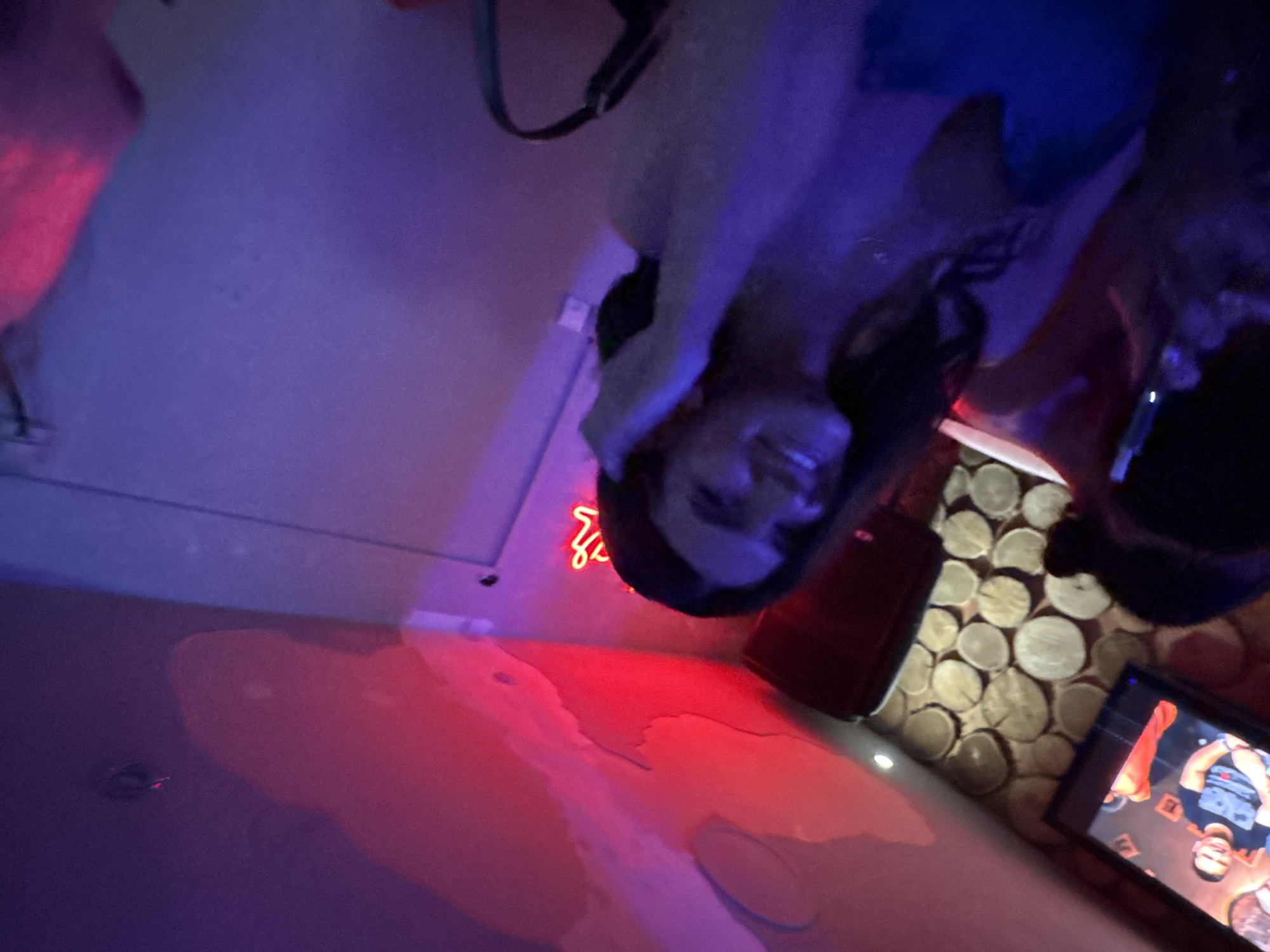 The image shows a room illuminated with pink and blue neon lights, possibly in Kragujevac, Serbia. A woman is visible in the scene, with dark hair, partially visible against a brightly lit background. In the foreground, there is a glossy surface reflecting the neon lights, suggesting a wet or polished surface. To the right, there is a table with a design featuring cross-sectional wood slices, and a mobile phone displaying a wrestling match. The overall scene suggests a modern, possibly nightlife-related environment.