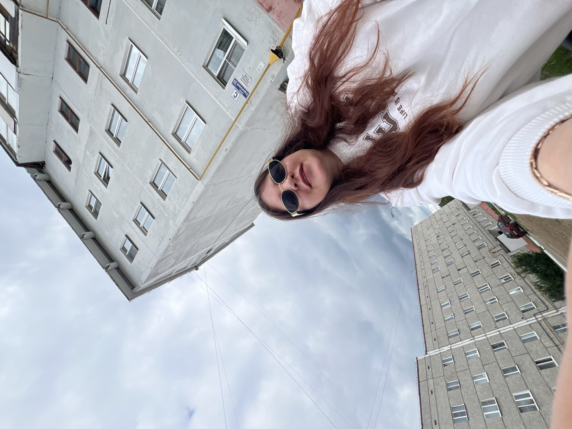 The image shows a young woman with long, reddish-brown hair and sunglasses, wearing a white sweatshirt with a graphic print. She is standing outdoors in front of a grey, multi-story apartment building. The building appears to be in a state of disrepair, with peeling paint and visible signs of aging. There are multiple windows on the building, some of which are open. The sky is overcast with dark clouds. In the background, another similar apartment building is visible, along with a tree and a red car. The scene is likely located in Noviy, Russia, given the architectural style of the buildings and the overall urban environment. The woman is looking directly at the camera, taking a selfie. There is no indication of any criminal activity or violence.