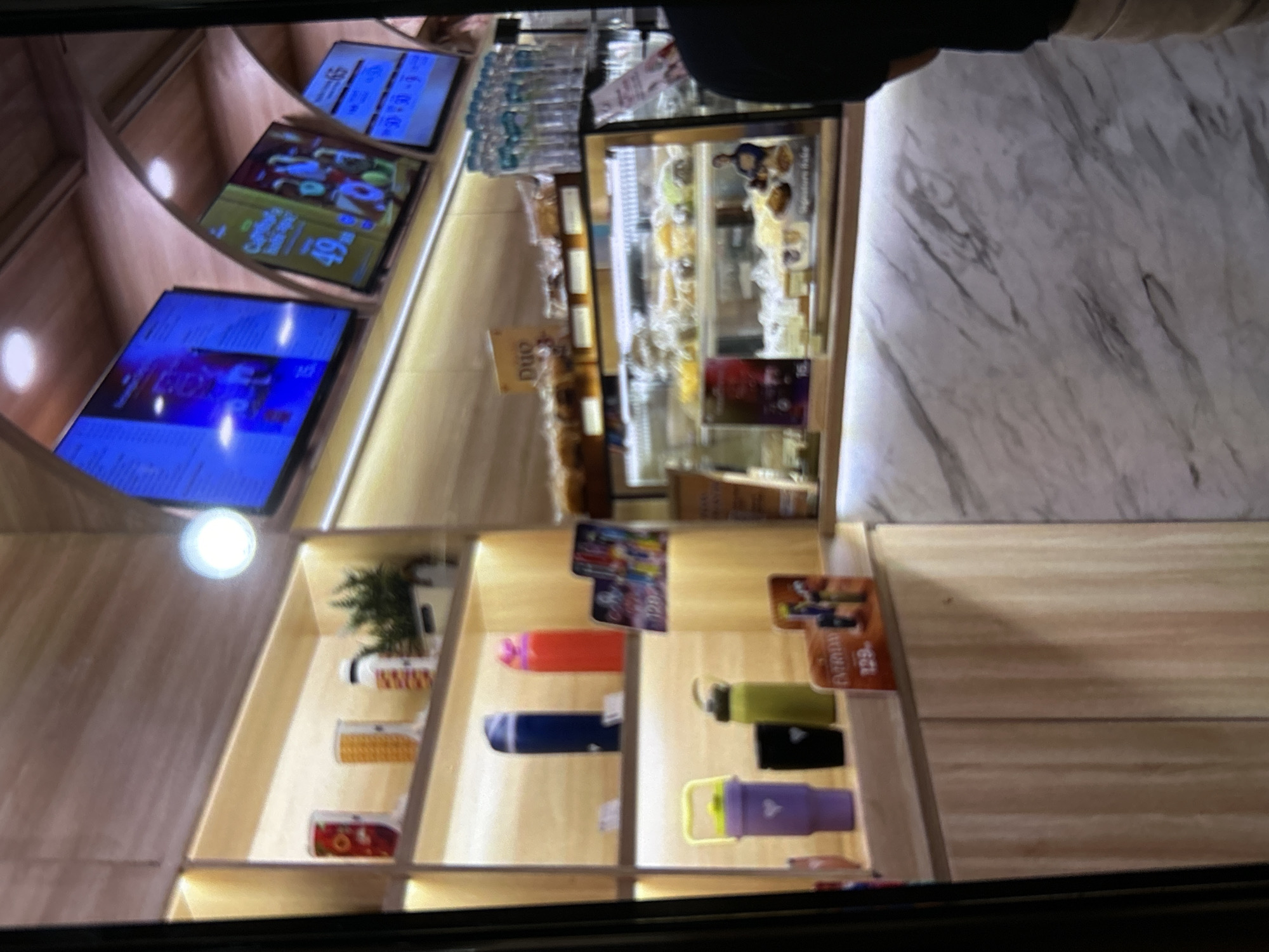 The image depicts the interior of a cafe or bakery in Depok, Indonesia. In the foreground, a person wearing a black t-shirt and light-colored pants stands partially visible on the right, facing towards the counter. The counter itself has a marble-like finish. Behind the counter, a display case filled with various pastries and baked goods is illuminated. Above the display case, shelves are stocked with water bottles and some decorative items. Further back, three digital screens are mounted on the wall, displaying menus or advertisements. The overall ambiance suggests a modern and clean establishment, likely a commercial space catering to customers seeking refreshments and baked goods.