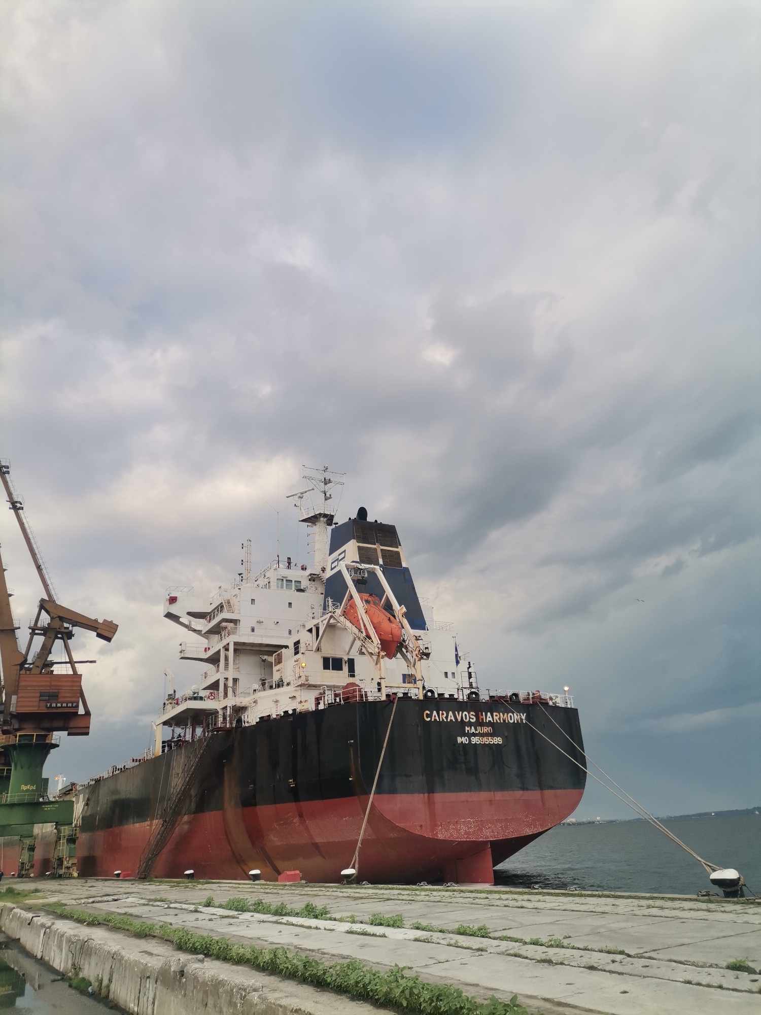 The image shows a large cargo ship, named "CARAVOS HARMONY" registered in Majuro (IMO 9595589), docked at a port. The ship's hull is black above the waterline and red below. It is secured to the dock with mooring lines. A large crane is visible on the left side of the ship. The dock is concrete with patches of vegetation growing on it. The sky is overcast. The image was taken in Ezerovo, Bulgaria.