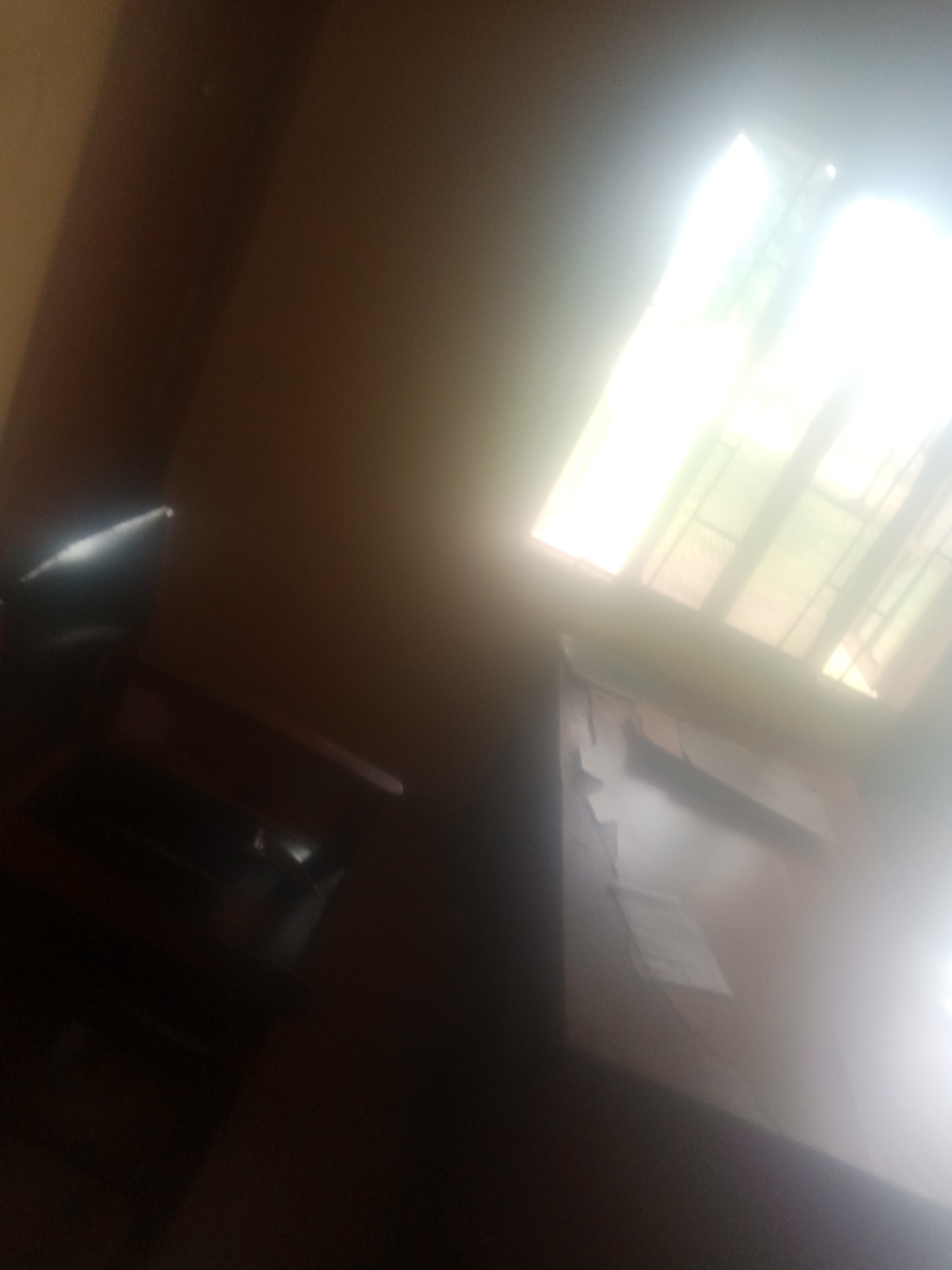 The image depicts an interior scene, likely an office or room, with a strong light source coming from a window or skylight. The lighting is very bright and overexposed in some areas, making it difficult to discern details.
In the foreground, a dark, possibly leather, chair is visible on the left side of the frame. To the right of the chair, a desk or table is partially visible, with some papers or documents on its surface. The rest of the room appears to be dimly lit or out of focus.
The prominent feature is the bright light from above, which suggests it is daytime, possibly with clear weather outside. The light is coming through a multi-paneled window or skylight, with grid-like structures visible within the light source. The overall impression is one of a somewhat sterile or functional space.
There is no visible text in the image. The scene is static, with no apparent activity taking place. The location context of Jalingo, Nigeria, does not provide specific visual cues within the image itself, as the interior is generic. The overexposed lighting creates a slightly abstract or ethereal quality rather than conveying specific emotions.