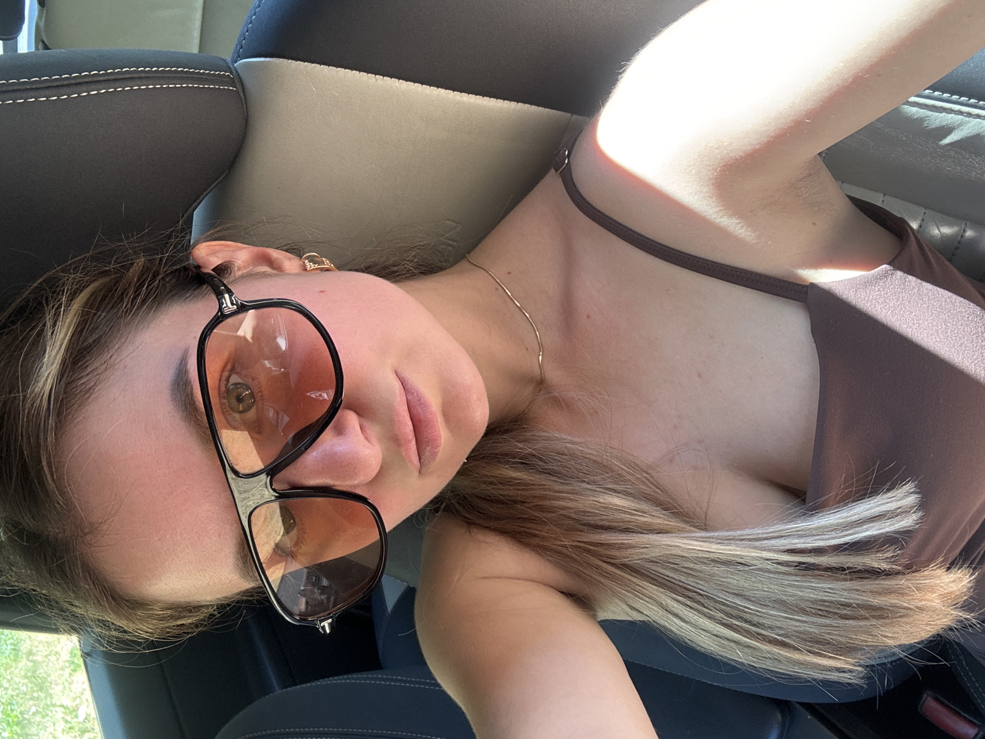 A young woman with blonde hair, wearing sunglasses and a brown tank top, sits in the passenger seat of a car. Her hair is pulled back and falls over her shoulder. She is wearing a thin gold chain necklace and a small gold earring in her left ear. The interior of the car is visible, with dark seats and a headrest. The background shows blurred greenery, suggesting an outdoor setting, possibly in Saint-Claude, France. The lighting indicates it is daytime.