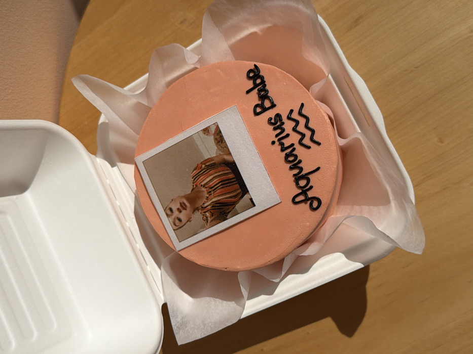The image displays a small, round cake with light peach-colored frosting, situated inside an open, white, rectangular molded-pulp take-out container. The container is lined with crumpled white tissue paper surrounding the cake. Centered on the cake's top surface is an edible image designed to mimic a Polaroid photograph. This image features a young adult female, depicted from the chest up, looking directly at the viewer with a slight head tilt. She is wearing a horizontally striped garment in orange, black, and white tones. Below this edible image, the words "Aquarius Babe" are inscribed in black icing, accompanied by two wavy lines, which represent the Aquarius zodiac symbol. The container rests on a light brown wooden surface. Distinct shadows are cast from the left side of the container onto the wooden surface, indicating a light source from the left. The scene is located in Tel-Aviv, Israel.