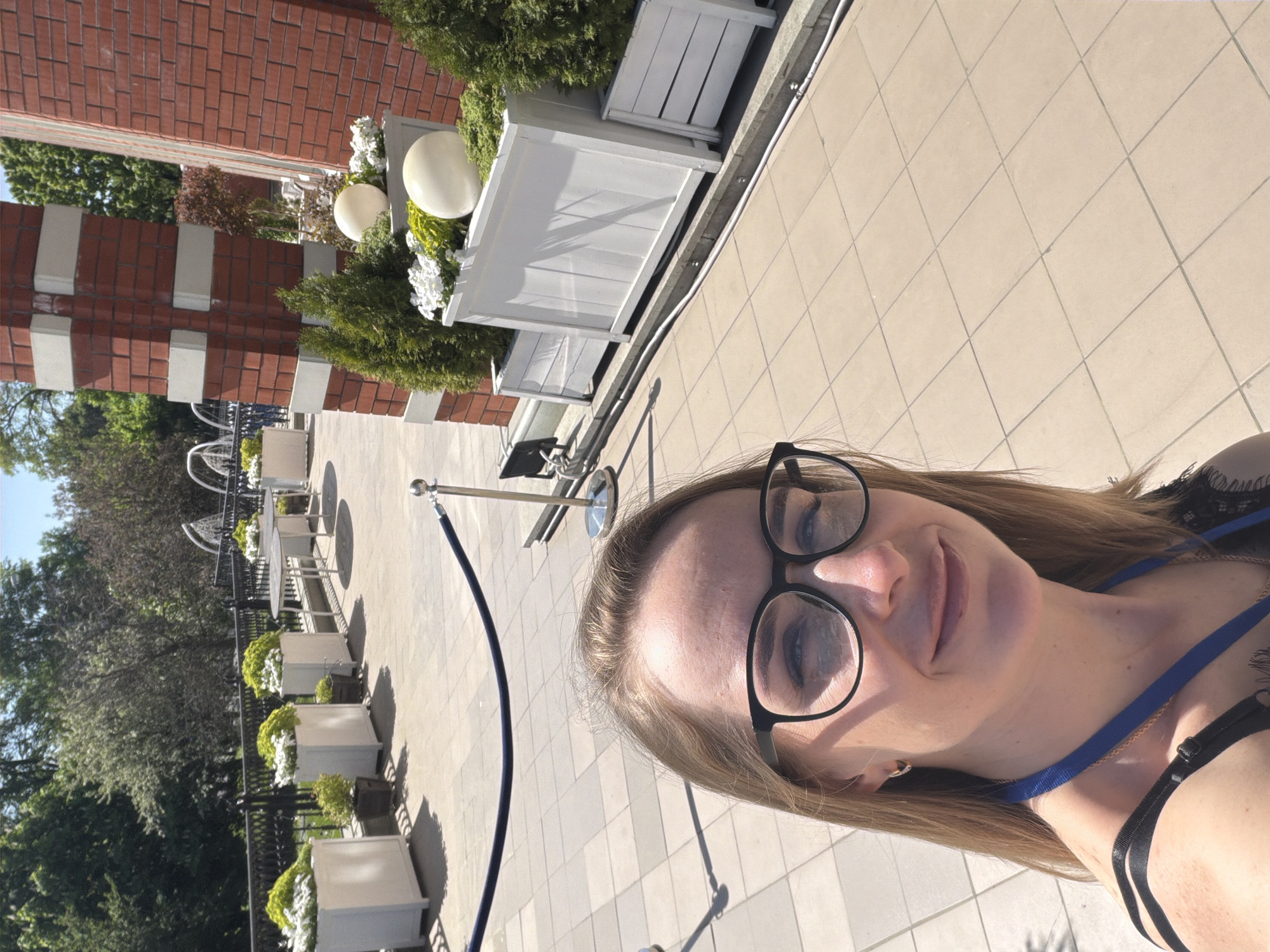 The image is a selfie of a woman wearing glasses, with a blue lanyard around her neck. She is standing outdoors in a public area with tiled paving. In the background are flower planters, some containing white flowers, arranged along a low wall. There are also round, white lamp fixtures. Further in the background, one can see tables and chairs with umbrellas, suggesting an outdoor seating area, possibly for a cafe or restaurant. A section of a brick building is visible on the left side. The overall setting suggests an outdoor space in a European-style urban environment. Based on contextual clues from the image, this is near the Gum shopping centre in Moscow, Russia.