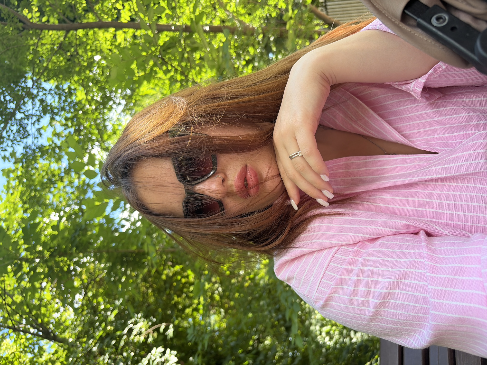 The image depicts a female individual, approximately 20-35 years old, positioned centrally and looking directly at the viewer. She has long, straight, reddish-brown hair, some strands of which are disheveled and fall across her face. She is wearing dark, oversized sunglasses that obscure her eyes. A silver-colored nose ring is visible on her right nostril. Her lips appear full, with a light pink or nude gloss. She is wearing a light pink collared shirt with thin white vertical stripes, unbuttoned at the collar to reveal a silver-colored necklace. Her right hand (viewer's left) is resting under her chin, with her fingers slightly curled. Her fingernails are manicured with a light, possibly white or off-white, polish. On her right ring finger, two rings are visible: a plain band and another with small stones.
The setting is outdoors on a sunny day, characterized by a dense background of vibrant green foliage from trees and bushes, indicating a park or garden environment. Sunlight filters through the leaves, creating numerous bokeh effects. A small patch of light blue sky is visible through the tree canopy in the upper-left quadrant. A dark wooden bench is partially visible behind the subject on the left. On the far right, a section of a beige baby stroller or pram, including part of its dark frame/handle, is visible. Based on the request, this scene is situated in the city of Krasnodar, Russia; however, no identifiable landmarks specific to Krasnodar are present in the image to independently confirm this location.