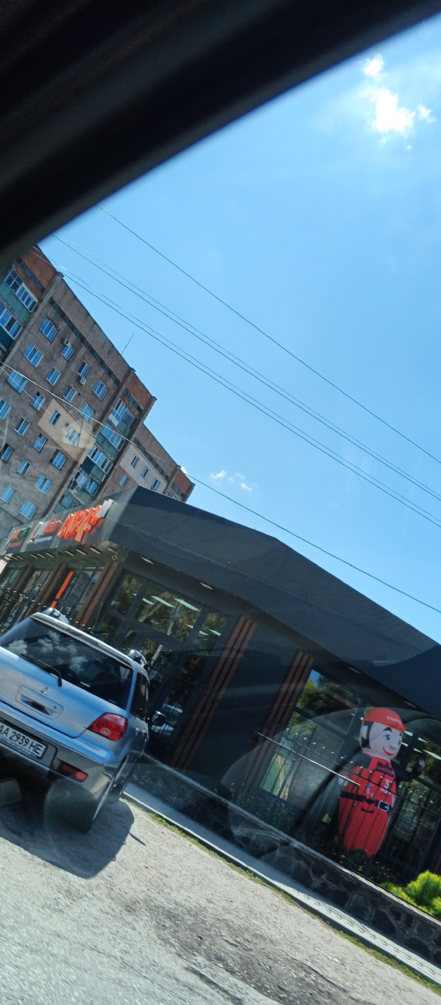 The image depicts a street scene in the city of Smila, Ukraine, on a clear, sunny day. In the foreground, a silver Mitsubishi Outlander SUV is parked on the asphalt road. The vehicle's license plate, partially visible, reads "AA 2939 HE". Behind the SUV, a modern building with a dark, sloped awning is visible. The building features large glass windows and appears to be a commercial establishment. An inflatable figure, resembling a cartoon character in a red outfit with a black helmet, stands near the entrance of the building, partially visible through the glass. In the background, a multi-story brick apartment building with numerous windows is seen against a bright blue sky with a few scattered clouds. Power lines are strung across the sky. The overall scene suggests an urban environment in Ukraine.