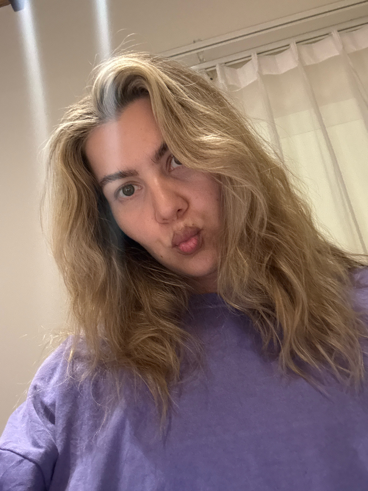 A blonde woman with light skin is posing for a selfie. She is wearing a purple t-shirt. Her lips are pursed as if blowing a kiss. Her hair is parted on the side and falls loosely around her shoulders. The background appears to be a room with a white curtain and some ambient lighting. There is no indication of location in the city of Unknown.