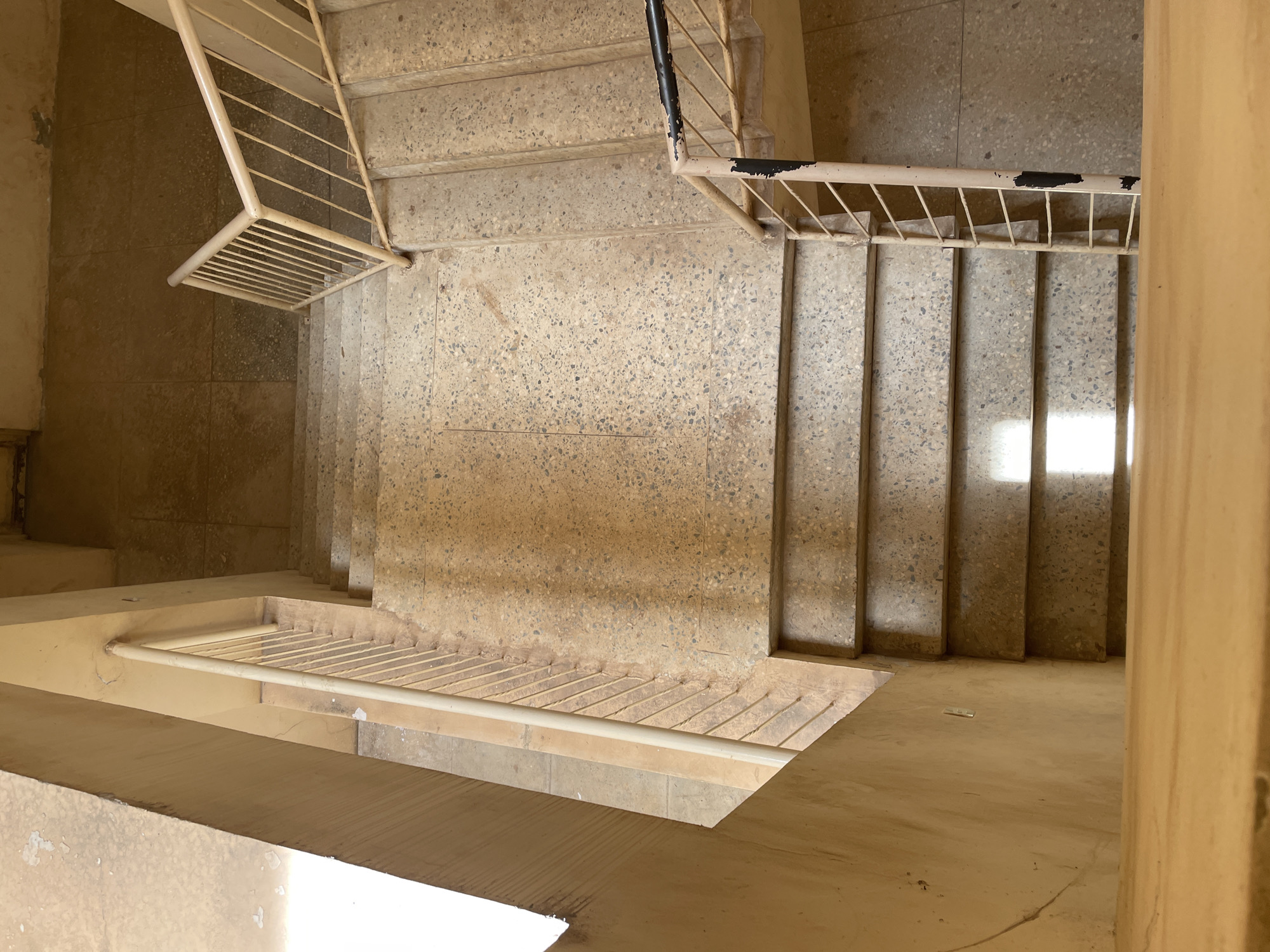 An overhead, full shot captures a concrete staircase with light beige railings. The staircase is sectioned into several tiers, with each step made of speckled beige concrete. A vertical railing, also light beige, runs along the left side of the stairs, with parallel metal bars connecting to the main banister. Sunlight illuminates a section of the steps, creating bright highlights. To the right, another staircase descends, with a similar beige railing that is chipped and has black paint patches near the bottom. The walls surrounding the staircase are a pale yellow, with some staining and marks visible. The overall scene appears to be an interior stairwell in a building, possibly in Kabala, Nigeria, given the texture and color palette often found in older structures in the region. There are no people or animals present in the image.