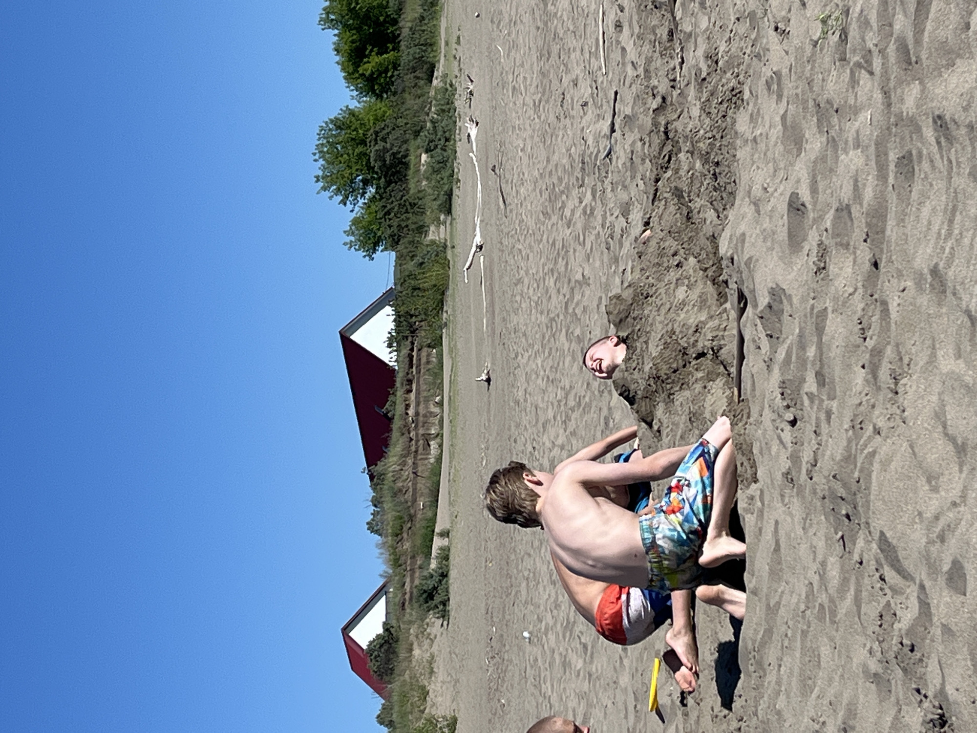 The image depicts a sunny day at a beach with several children playing in the sand. In the foreground, two boys are kneeling near a third boy who is buried in the sand, with only his head visible and his mouth open in laughter. The boy in the middle is wearing patterned swim trunks, and the boy to his left is wearing red and white striped swim trunks. A yellow shovel lies in the sand to the left. In the background, two houses with red roofs are visible on a sandy dune dotted with green vegetation. The sky is clear and blue, and driftwood is scattered across the sand. The scene suggests a recreational activity at a beachside location. There is no information available to place this scene in Krasnoturansk, Russia.