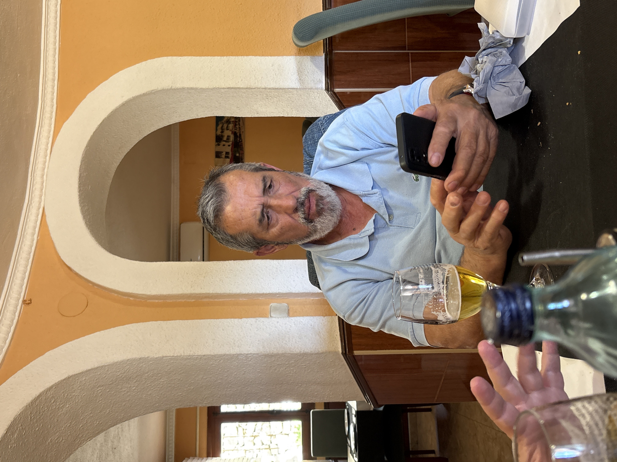 A man with graying hair and beard sits at a table in a restaurant, holding a smartphone. A glass of beer sits to his left, and a bottle of water is in the foreground. The table is covered with a black tablecloth. The restaurant features arched walls painted in a combination of orange and white. The setting is likely in Teulada, Spain.