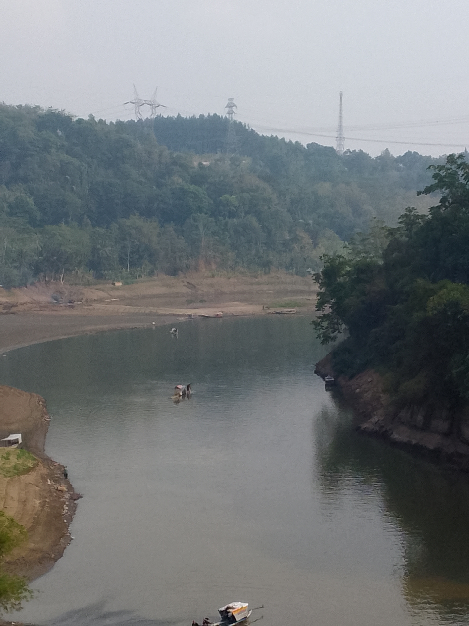 A hazy, overcast day looms over a wide, calm river bordered by steep, tree-covered hills. The riverbank on the left is steep and appears to be composed of dried mud, with a small white structure perched on top. A few small, wooden boats are scattered across the water. One boat in the foreground carries three figures, appearing to be working or fishing. On the far bank, dense green foliage covers the hilly terrain, with power lines and transmission towers visible in the distance against the pale sky. The overall atmosphere is one of quiet solitude and natural beauty, with a touch of human presence.