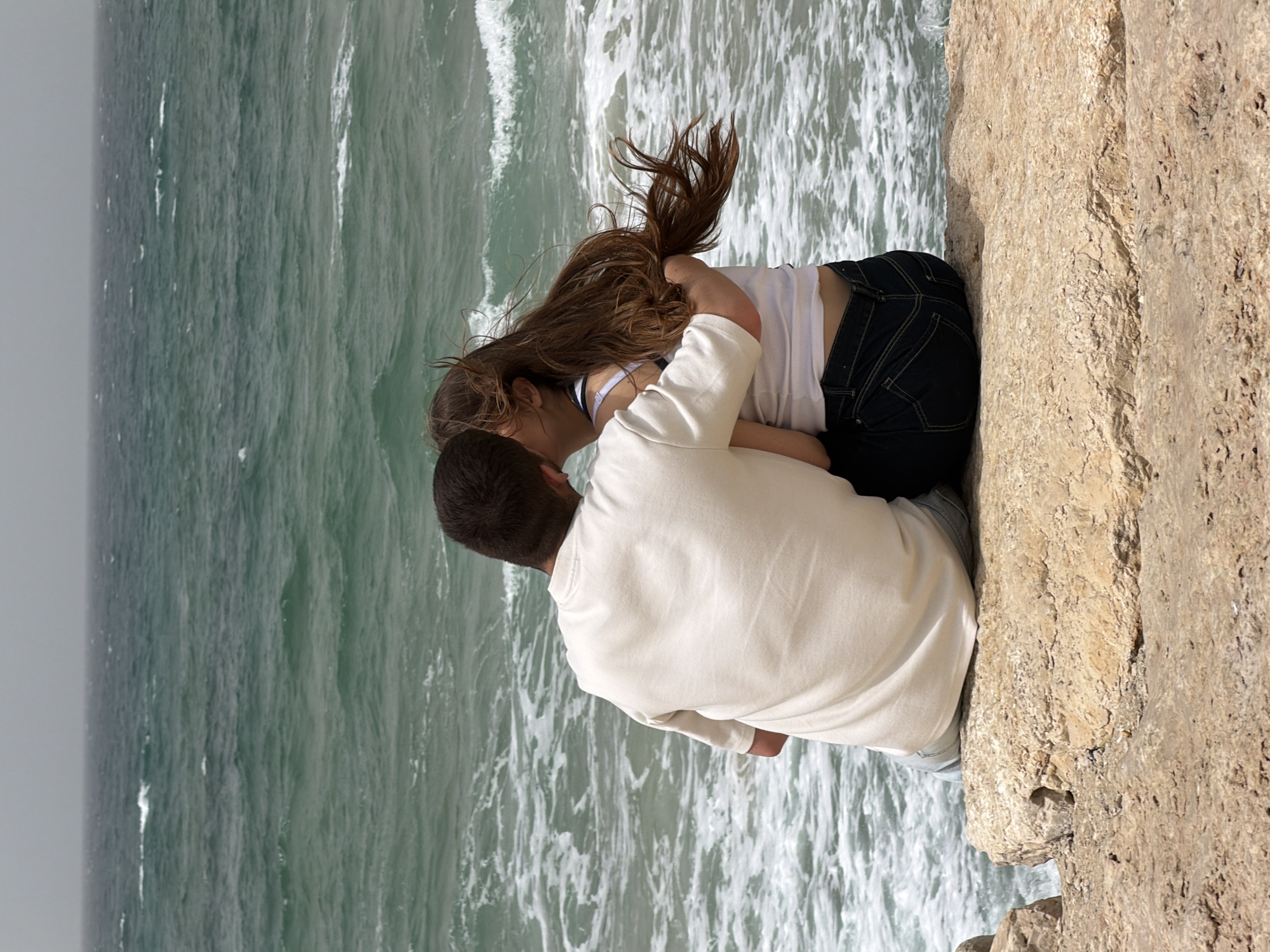 A man and woman embrace on a cliff overlooking the Mediterranean Sea. They appear to be on a cliffside overlooking the sea. The water below is turbulent with waves. The cliff is composed of sedimentary rock. The location is likely near Tel Aviv, Israel, given the coastal environment. The man wears a light-colored shirt and lighter pants, and the woman wears a white shirt and dark jeans. Her hair is loose.