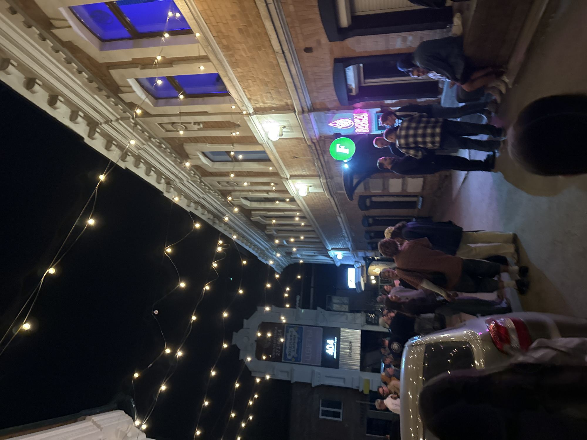 The image shows a nighttime street scene in Krasnoyarsk, Russia. String lights are strung overhead along a building facade with illuminated skylights. People are gathered on the sidewalk, some standing in front of a bar or establishment with a neon sign. The sign of the bar has the name "Brakhauz Brauerei." A car is parked on the street with its taillights visible. A sign with the number "404" is visible in the background.