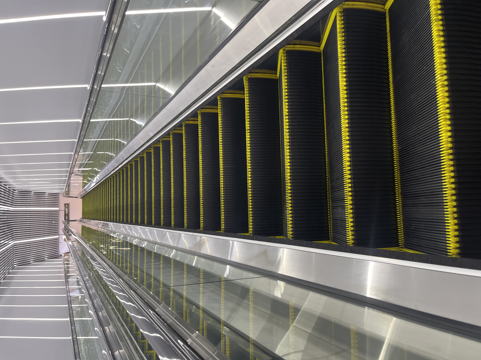 An upward-facing, wide-angle shot shows a modern escalator at an unknown location in Shapingba District, China. The escalator has black steps with yellow safety strips. Clear glass railings flank both sides, reflecting the bright, vertical white lights that line the walls and ceiling of the structure. The ceiling features a striped, metallic design. The escalator appears to be empty.