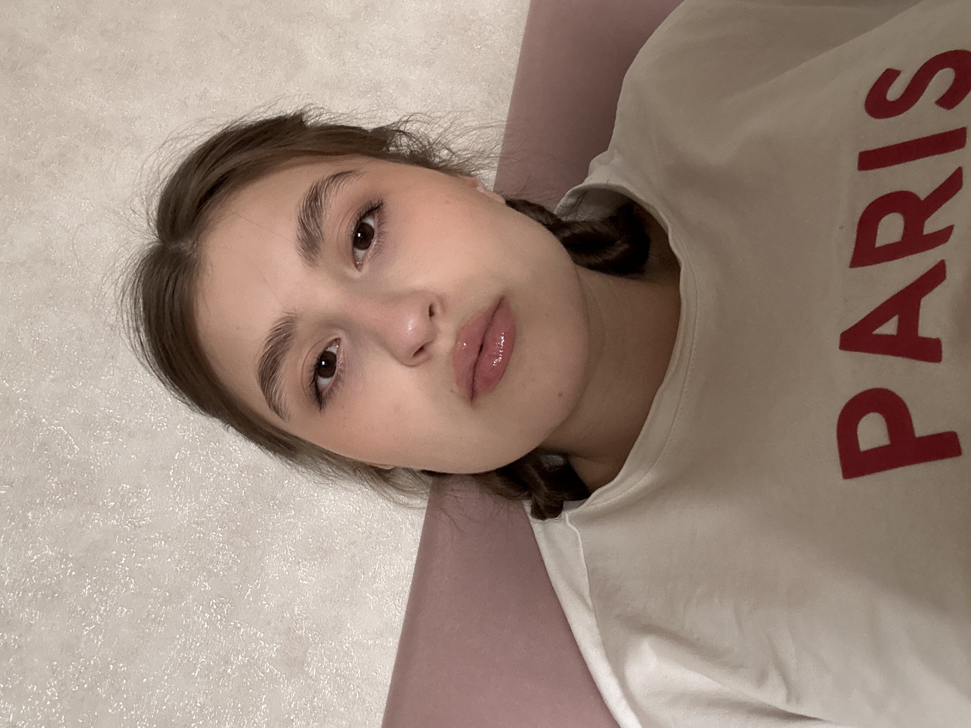 The image displays a young female individual situated indoors in Sarapul, Russia. The subject has medium-brown hair, with two braids visible on either side, and dark brown eyes looking directly forward. Her lips are full and appear to have a glossy application. She is wearing a light-colored, possibly off-white or beige, crew-neck garment featuring the word "PARIS" in red capital letters across the chest. The immediate background consists of a textured light-colored wall with a subtle pattern and a soft pink surface, possibly a pillow or furniture armrest, to the viewer's left. The subject is in a static pose, looking towards the camera.