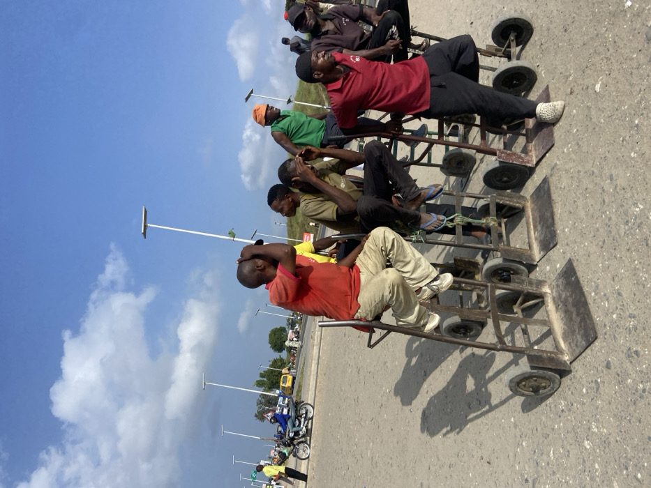 The image depicts an outdoor scene featuring several men seated on custom-built, metal-framed trolleys on what appears to be an asphalt or concrete road. There are at least six adult males visible in various colored shirts (red, green, yellow, brown) and long pants, some wearing sandals or shoes.
The trolleys are robust, multi-wheeled structures, seemingly designed for sitting, or potentially for transporting goods. They cast distinct shadows on the ground, indicating a bright, sunny day. The men's postures vary; some look forward, others down or to the side. One man in the foreground, wearing a red shirt, has his hands clasped on his head, while another in a brown shirt appears to be gesturing with his hands.
In the background, under a clear blue sky with scattered white clouds, modern white streetlights or poles line the road. Further down, several motorcycles and other vehicles are partially visible, suggesting an urban or semi-urban setting consistent with Dar es-Salaam, Tanzania. The overall impression is one of men resting or waiting, possibly involved in informal transport services using these trolleys. There is no discernible text visible in the image.