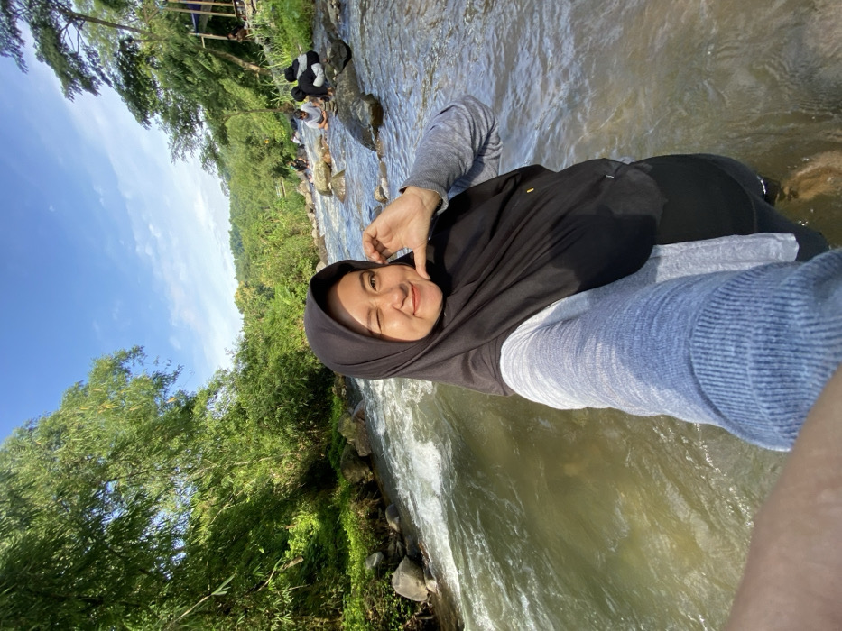 A woman in a black hijab and gray shirt stands in a river. Lush green trees and a blue sky with a few clouds form the backdrop. People are seen along the riverbank in the distance. The water flows around her legs. She is smiling and winking, holding up two fingers in a peace sign. The photo is taken at an angle, making the scene slightly tilted.