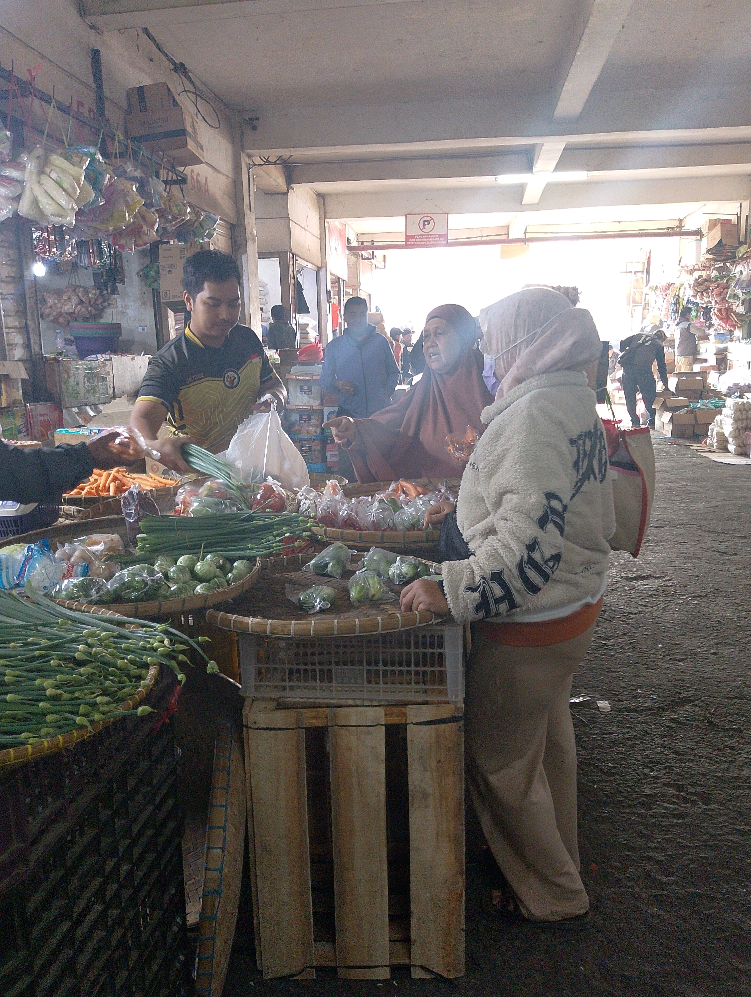 This is a medium shot, taken from a slightly low angle, of a bustling market stall in Lembang, Indonesia. Sunlight streams in from the right, creating bright highlights and deep shadows. The scene is filled with produce. In the foreground, a man in a yellow and black shirt is handing a plastic bag of vegetables to a customer. In front of him are baskets overflowing with green onions, small green gourds, and red chilies. To the right, a woman wearing a light-colored, fuzzy jacket and a pink headscarf stands with her back to the camera, appearing to be looking at the produce. Behind her, another woman in a dark maroon headscarf is visible, and beyond them, a crowd of people can be seen milling about. The market appears to be under a covered structure with a concrete ceiling and exposed beams, and the ground is a rough, dark surface. Various bags of produce and other goods are displayed on shelves and tables in the background, suggesting a vibrant and active marketplace.