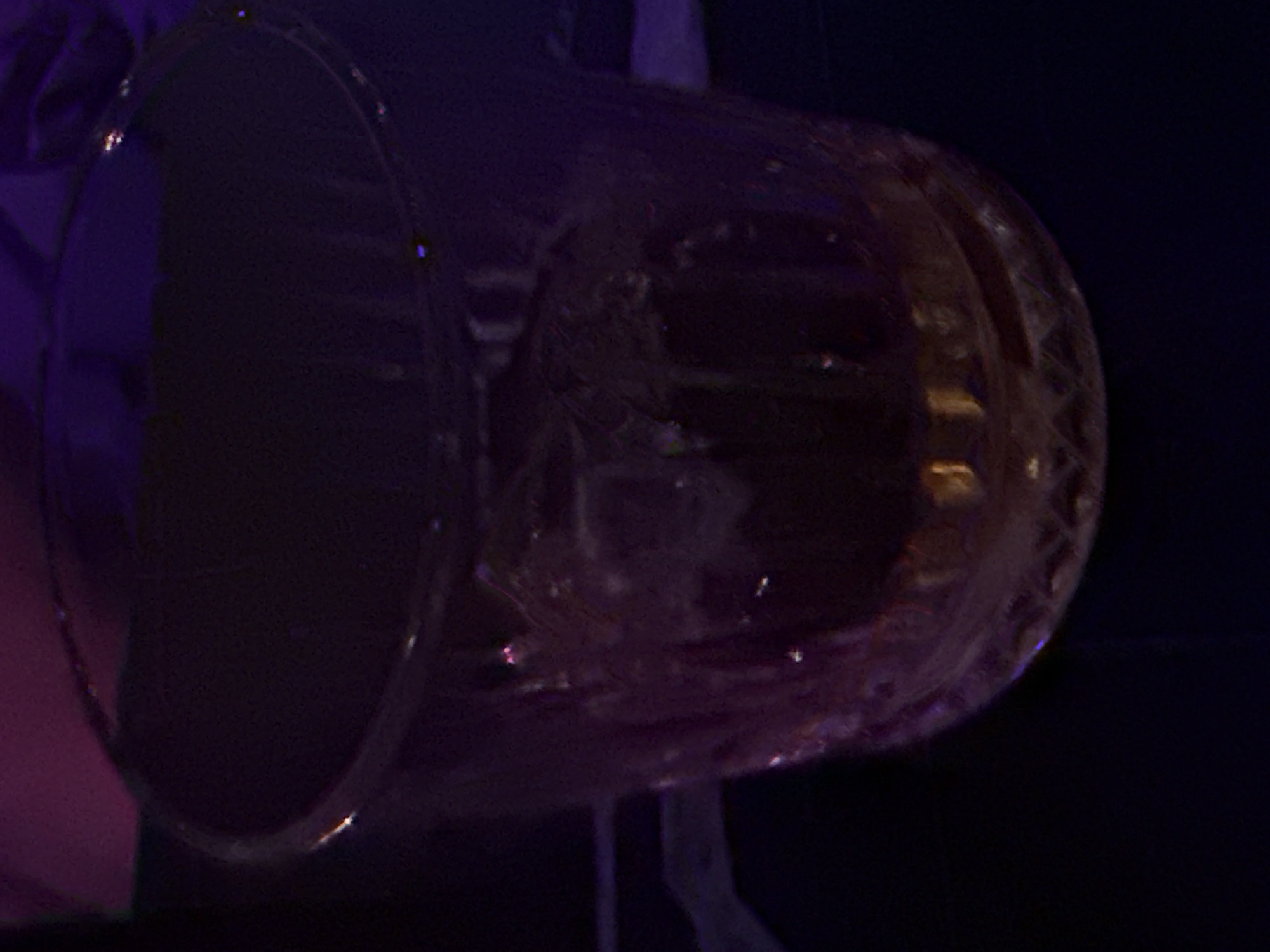 A clear, short tumbler glass, featuring a patterned base, is centrally depicted, containing an amber-colored liquid and multiple ice cubes. The glass rests on an unidentifiable dark surface. The environment is characterized by extremely low light conditions, with diffused purple and pink ambient illumination reflecting off the glass and softly illuminating the blurred background. No individuals are present or discernable. There are no visible actions or interactions. The setting appears to be an indoor, atmospheric venue, such as a bar or lounge. No identifying architectural features, signage, or other contextual elements are present to determine a specific geographic location, including the city of Sofia, Bulgaria.