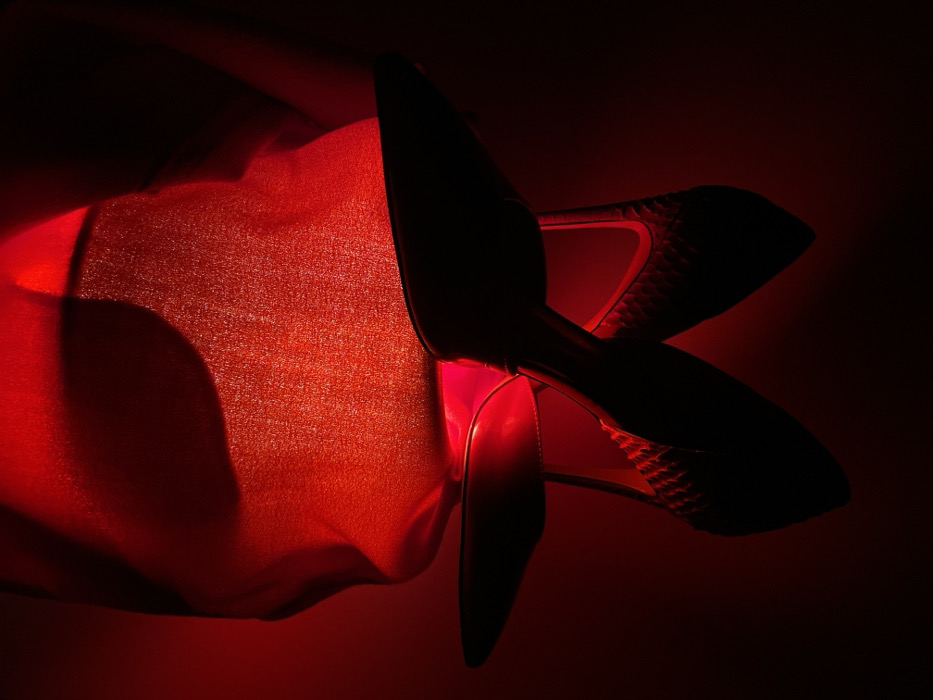 A high-angle shot under red lighting reveals a pair of high heels resting on a textured red fabric. The shoes are black, their pointed toes in sharp contrast to the fabric's soft texture. A hint of snakeskin pattern appears on the heel cups. The lighting emphasizes the shapes and textures, creating a dramatic effect with deep shadows and bright highlights.