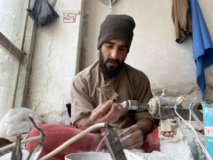 Within a modest workshop located in Kandahar, Afghanistan, a male artisan with a dark beard, mustache, and wearing a dark beanie and light brown tunic, is depicted from the chest up, intently focused on his work. His hands, dusted with white powder, are precisely manipulating a small, white, cylindrical object against a rotating spindle of a tabletop grinding or polishing machine. This machine, showing exposed electrical wires (green, pink, white) and covered in white powder, sits on a surface partially obscured by a red fabric draped over the artisan's lap. The immediate foreground contains blurred metallic tools and fragments. The background features unpainted, cracked plaster walls. To the left, a window frame allows natural light, and a black plastic bag hangs near a small white sign with Arabic script, likely "Ya Allah". To the upper right, a blue fabric and a brown garment hang from the wall. The scene is characterized by accumulated fine white powder, indicating ongoing material processing.