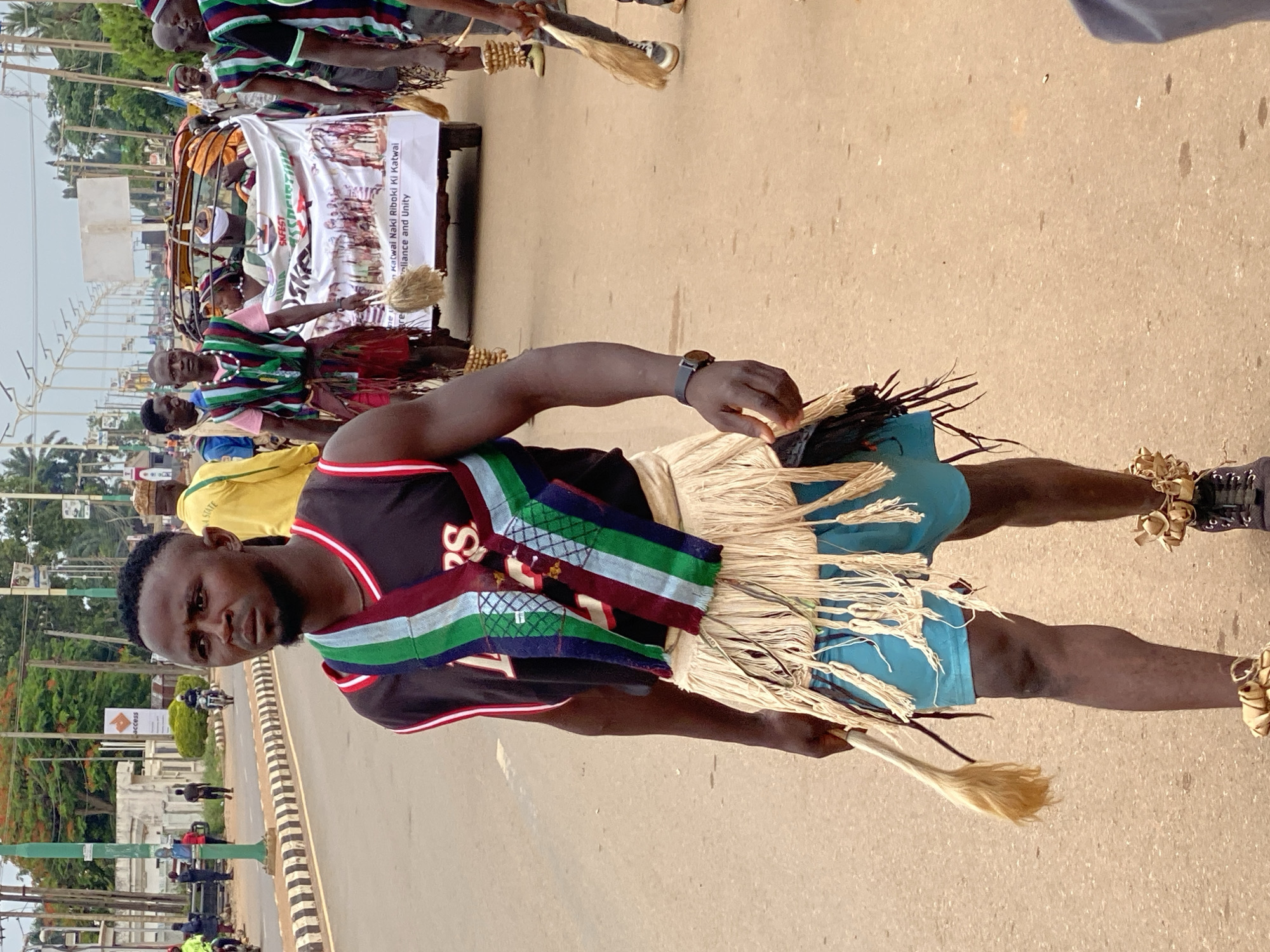 Here's a detailed description of the image:
**Scene:** A daytime outdoor setting on what appears to be a city street. The photograph is taken at ground level and is rotated, such that elements appear sideways.
**Subject:** The foreground features a man of African descent. He wears a black basketball jersey, blue shorts, and a straw-like fringed skirt or apron. A multi-colored (red, green, blue) sash is draped over his shoulder. He is holding a whisk broom, one in each hand. There are ankle rattles on his legs.
**People:** Other individuals are visible in the background, some wearing similar striped clothing and straw hats. There is a banner, although the text is partially obscured.
**Objects:** Besides the clothing and accessories mentioned above, there is a cart behind the main subject. The banner on the cart displays text and images; words like "Katwal Naki Riboki Ki Katwal," "Reliance and Unity," and "SKFEST" are visible, along with illustrations of people. Other objects include various traffic poles.
**Actions/Interactions:** The man is standing on the street and is looking directly toward the camera. It appears as though the people in the background are involved in a parade or procession of some kind.
**Environment:** The setting is a city street, likely in Nigeria, based on the attire and the "Access" sign. There are trees, buildings, and poles visible in the background. The street appears to be paved.