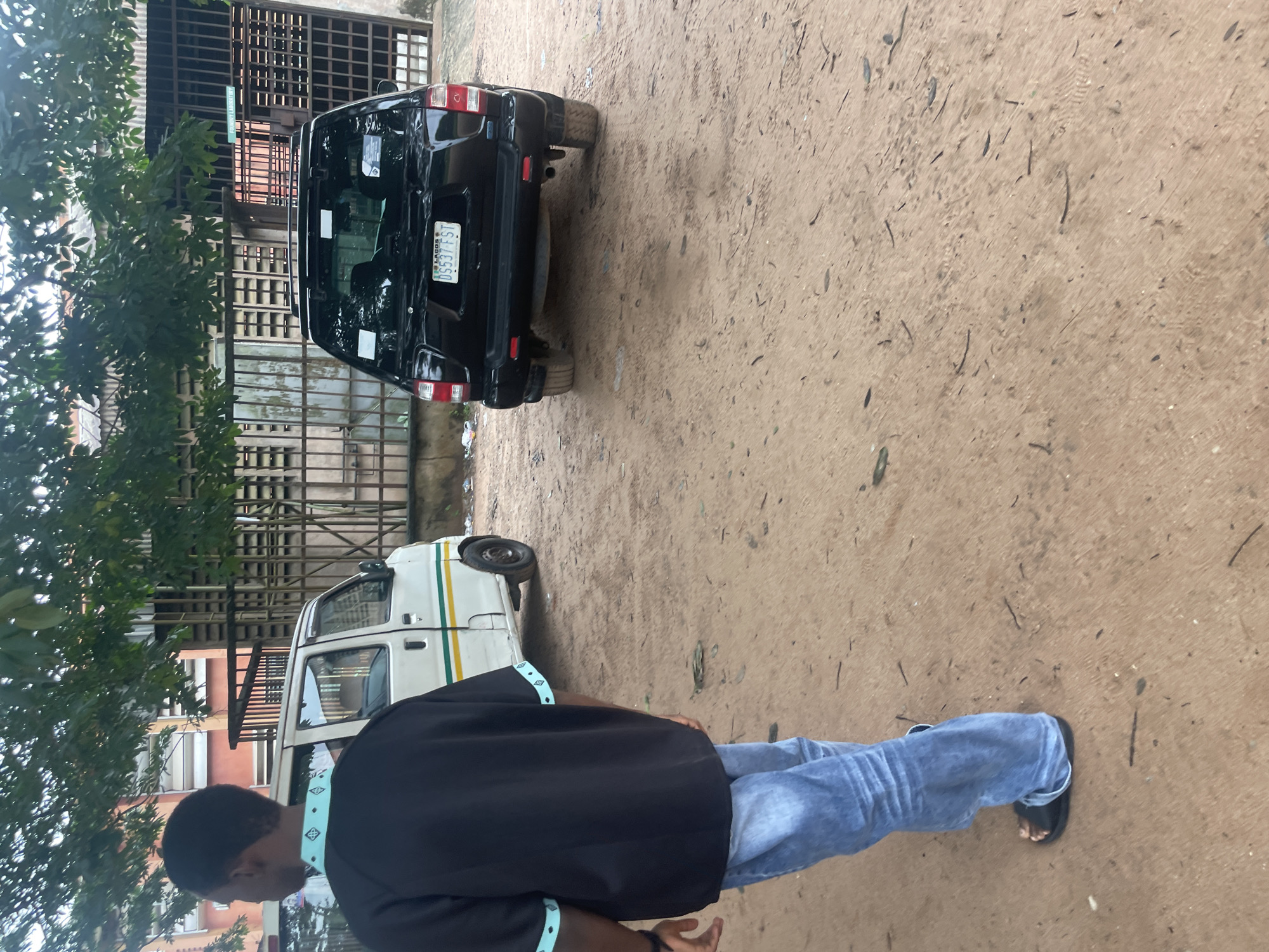 In Ihiagwa, Nigeria, a person in a black shirt with light blue trim and blue jeans, wearing black slides, walks away from the camera on a dirt ground. To the left, a white van with green and yellow stripes is parked. To the right, a black SUV faces away from the camera, displaying a Nigerian license plate that reads "DS537 FST". Behind the vehicles is a metallic fence and a building with windows, partially obscured by trees. The ground is sandy and littered with small debris.