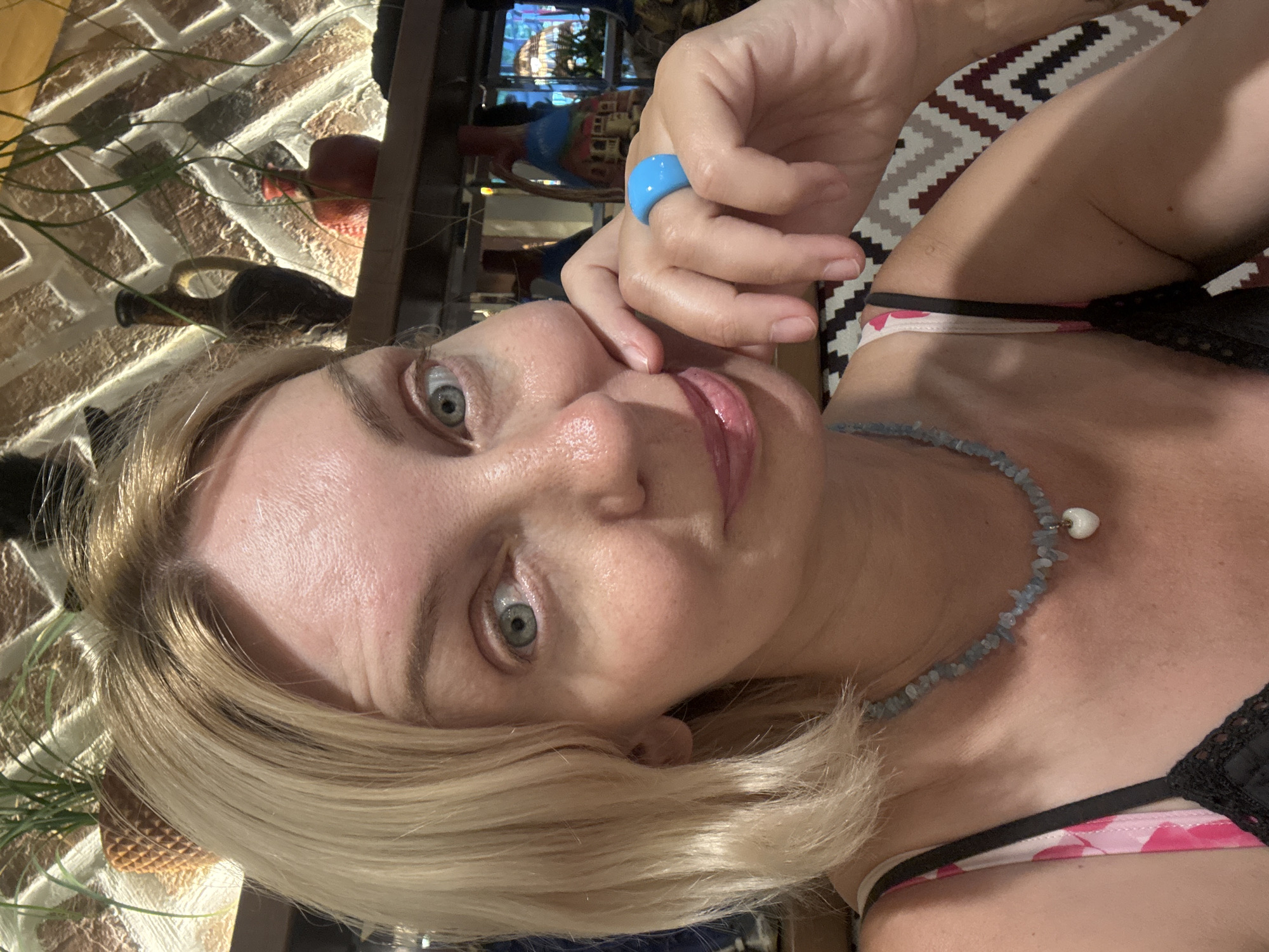 This is a close-up, low-angle selfie of a blonde woman in what appears to be an indoor setting with some decorative elements. The woman is looking directly at the camera with a neutral expression. She has fair skin, light blue eyes, and her blonde hair is styled in a bob cut. She is wearing a light blue ring on her right index finger and a necklace made of small blue stones and a single white pearl pendant. She is also wearing a low-cut top with a floral pattern, and a black lace bra is visible underneath.
The background is blurred but suggests a dimly lit room with textured walls, possibly brick. There are some indistinct objects in the background, including what looks like a vase and some greenery. A patterned textile is visible on the right side of the image.
The overall impression is a casual, personal photograph taken indoors, likely in a home or restaurant environment. There is no visible text in the image. The lighting suggests it could be evening or a dimly lit indoor space.