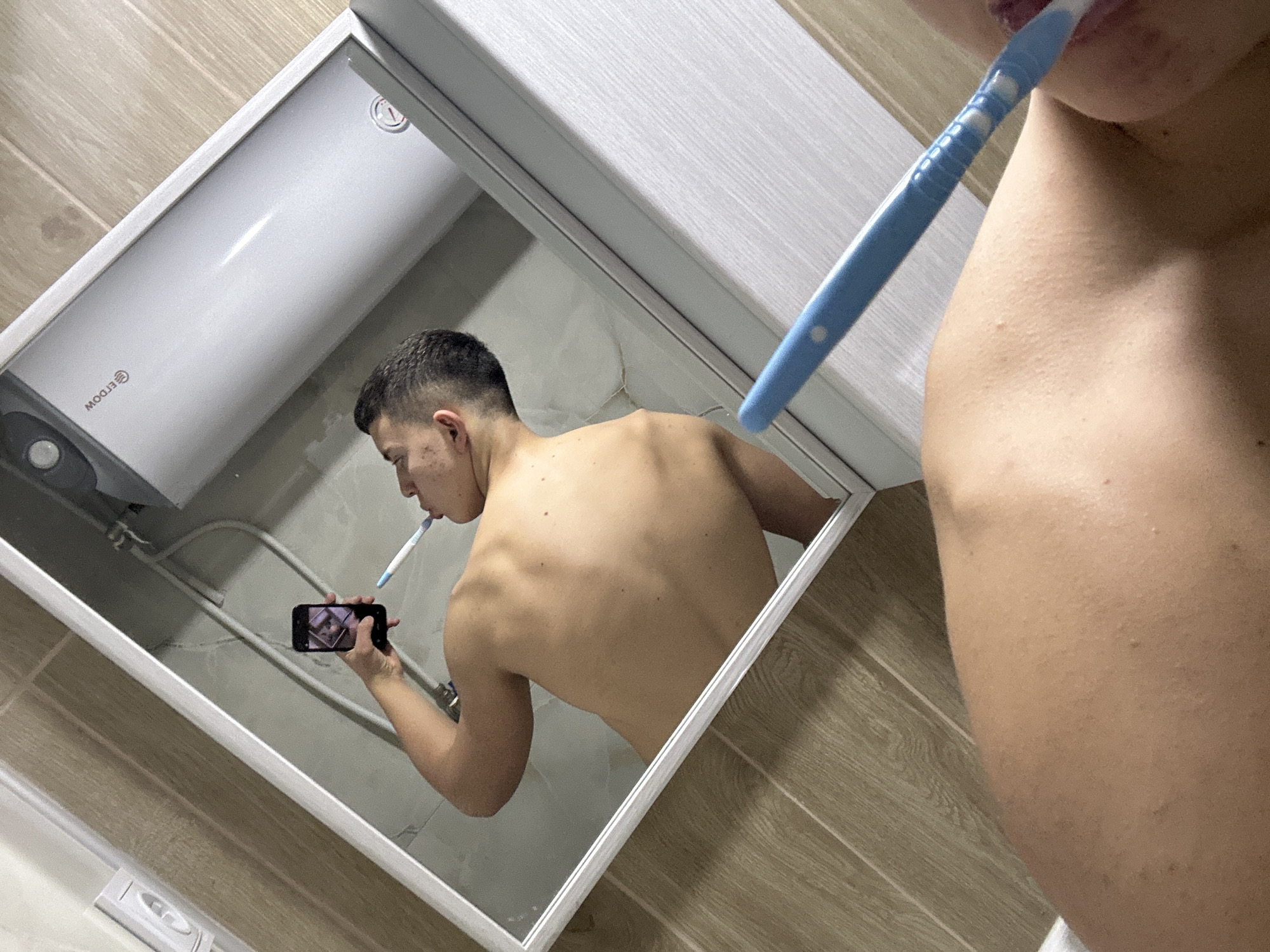 A shirtless man is brushing his teeth while looking at his phone in a bathroom mirror. The mirror reflects a water heater on the wall. The bathroom has wood-look tile. The location is presumed to be Zlatograd, Bulgaria.