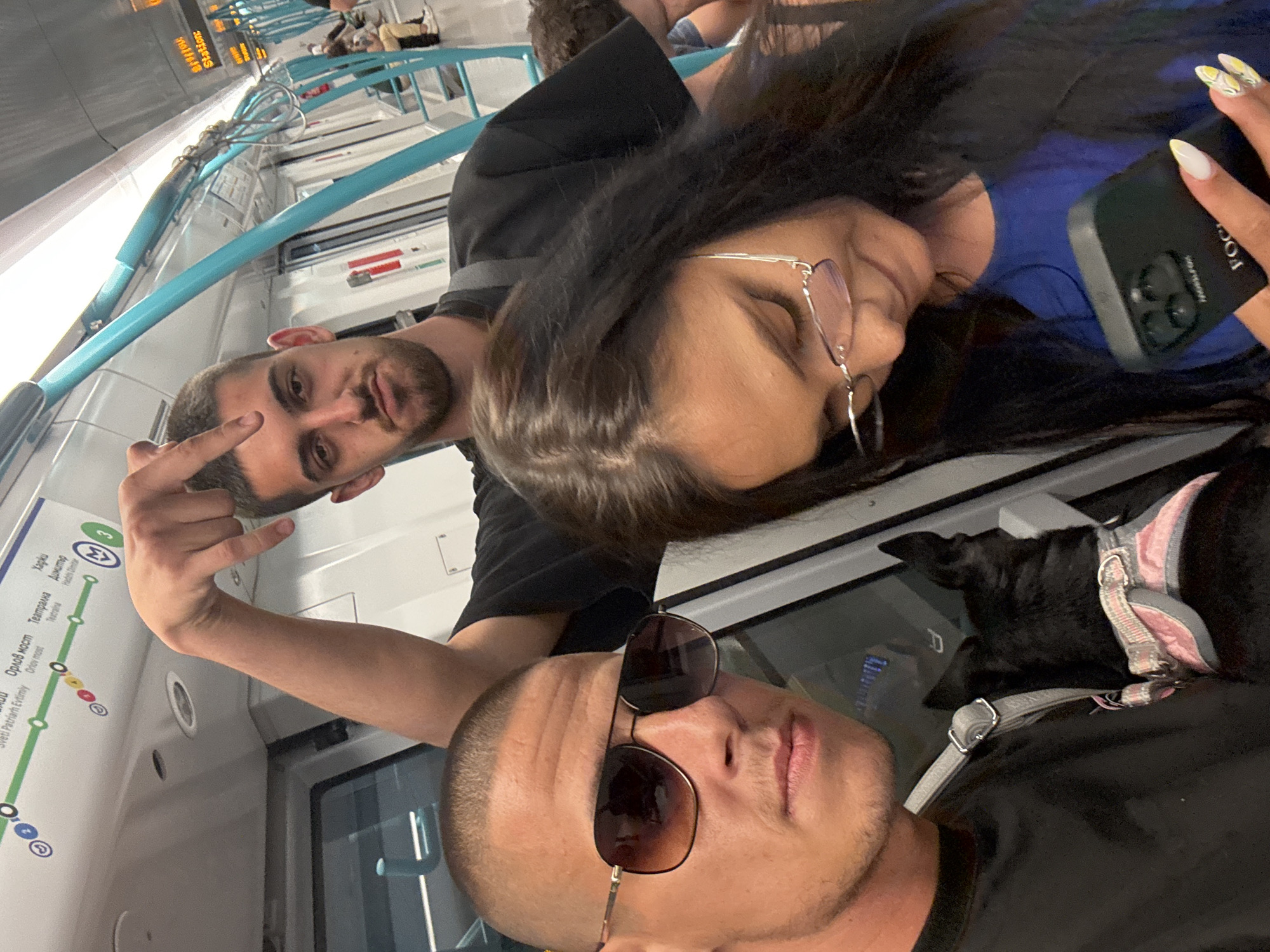 Three people and a dog are inside a Sofia Metro train. The person in the foreground on the left is a man with a shaved head wearing sunglasses and a black t-shirt. He has a black dog with a pink harness resting on his shoulder. To his right is a woman wearing glasses and a blue shirt, looking down at her phone. Behind them, a man with short dark hair and a beard is leaning forward, pointing downwards with his right index finger. The train interior shows teal-colored poles and overhead signage indicating metro stations.