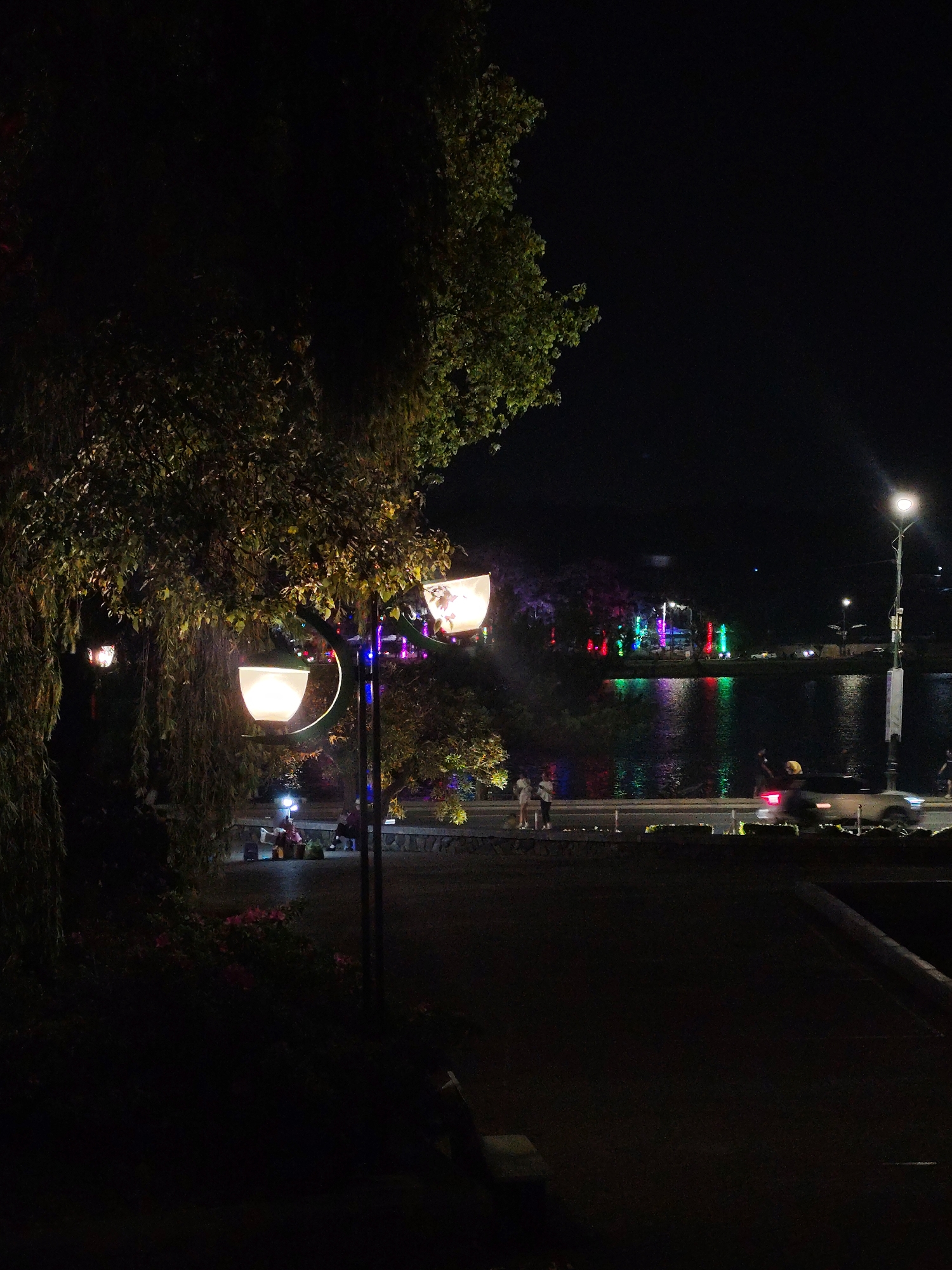 The image captures a night scene at Xuan Huong Lake in Da Lat, Vietnam. In the foreground, a lit double-lamp street light illuminates a park area with a weeping willow tree on the left and flowers in a garden bed. Further back, several people are sitting near another street light. Across the street, individuals stroll near the lake's edge, where a car's lights create a blurred streak as it passes. On the opposite side of the lake, colorful lights are visible, reflecting off the water's surface, indicating decorative installations or buildings. A taller street light stands to the right of the lake, adding to the scene's illumination. The environment appears dark, with details mainly visible where light sources illuminate the surrounding area.