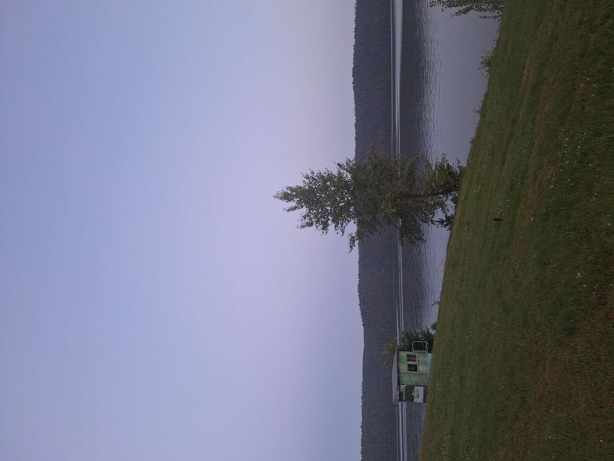 A wide-angle, low-light, outdoor, eye-level shot captures a serene natural landscape. The scene is dominated by a body of water, likely a lake or river, reflecting the soft hues of a twilight sky. The sky itself is a gradient of pale blue and lavender, suggesting either dawn or dusk.
In the foreground, a grassy slope slopes down towards the water. The grass is green and appears somewhat unkempt, with scattered white seed heads visible. A small, weathered, light green structure, resembling a small cabin or shed with a peaked roof and windows, is situated on the left side of the slope. Next to this structure, a young tree with sparse foliage is visible.
Further back, a larger tree with a fuller canopy stands to the right of the cabin. Its leaves are a deep green, providing a contrast to the lighter tones of the grass and sky. The middle ground is occupied by the body of water, its surface rippled by a gentle breeze.
Beyond the water, a dense line of dark green trees forms a treeline along a distant, undulating hillside. The foliage of these trees is so thick that individual trees are indistinguishable, creating a dark, textured mass against the lighter sky. The overall impression is one of quiet solitude and natural beauty, possibly in a rural or park-like setting. The location is not specified in the image or the provided hints.