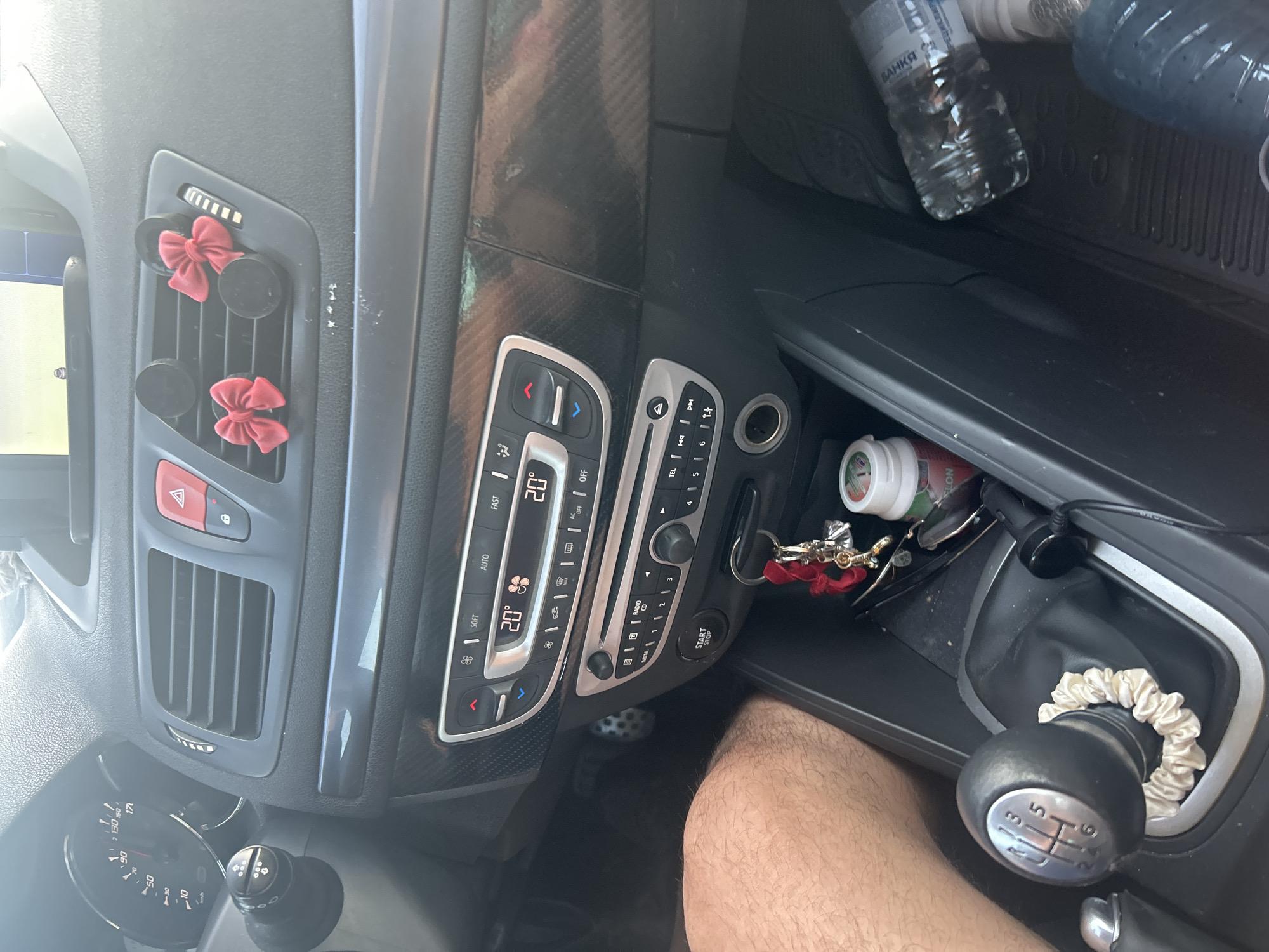 The image shows the interior of a car, likely a hatchback, parked on a street. The dashboard features a digital display showing "20°" on both sides, indicating the air conditioning settings. There are ventilation grilles, a hazard light button, and decorative red bows attached to one of the grilles. A manual gear stick with a fabric scrunchie on its base is visible, along with a key holder with red charms. Below the gear stick, a "START STOP" button and a power outlet are present. On the passenger side, a plastic water bottle is placed on the floor mat. The image is taken from the driver's perspective, showing the dashboard, center console, and part of the passenger seat. The car appears to be parked in a sunny outdoor environment. Based on the provided context, the location is identified as Byala Slatina, Bulgaria.
