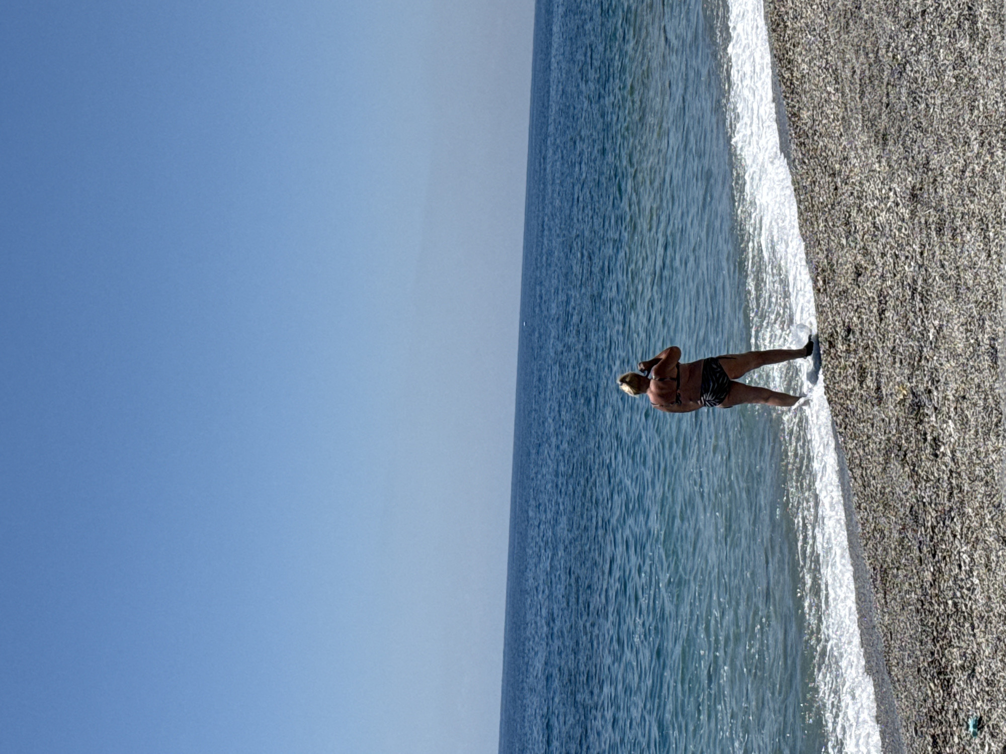 A slightly rotated outdoor shot shows a woman standing in the surf. The scene is dominated by a clear blue sky occupying most of the left side of the image. To the right, the blue sea stretches to the horizon. The edge of the sea meets a pebble beach where a woman is standing. She is facing away from the camera, wearing dark swimwear, and has short hair. Her arms are bent, possibly adjusting something on her head or neck. The shoreline is defined by the white foam of gentle waves lapping against the dark pebbles of the beach. The location of the image is Unknown, Unknown.