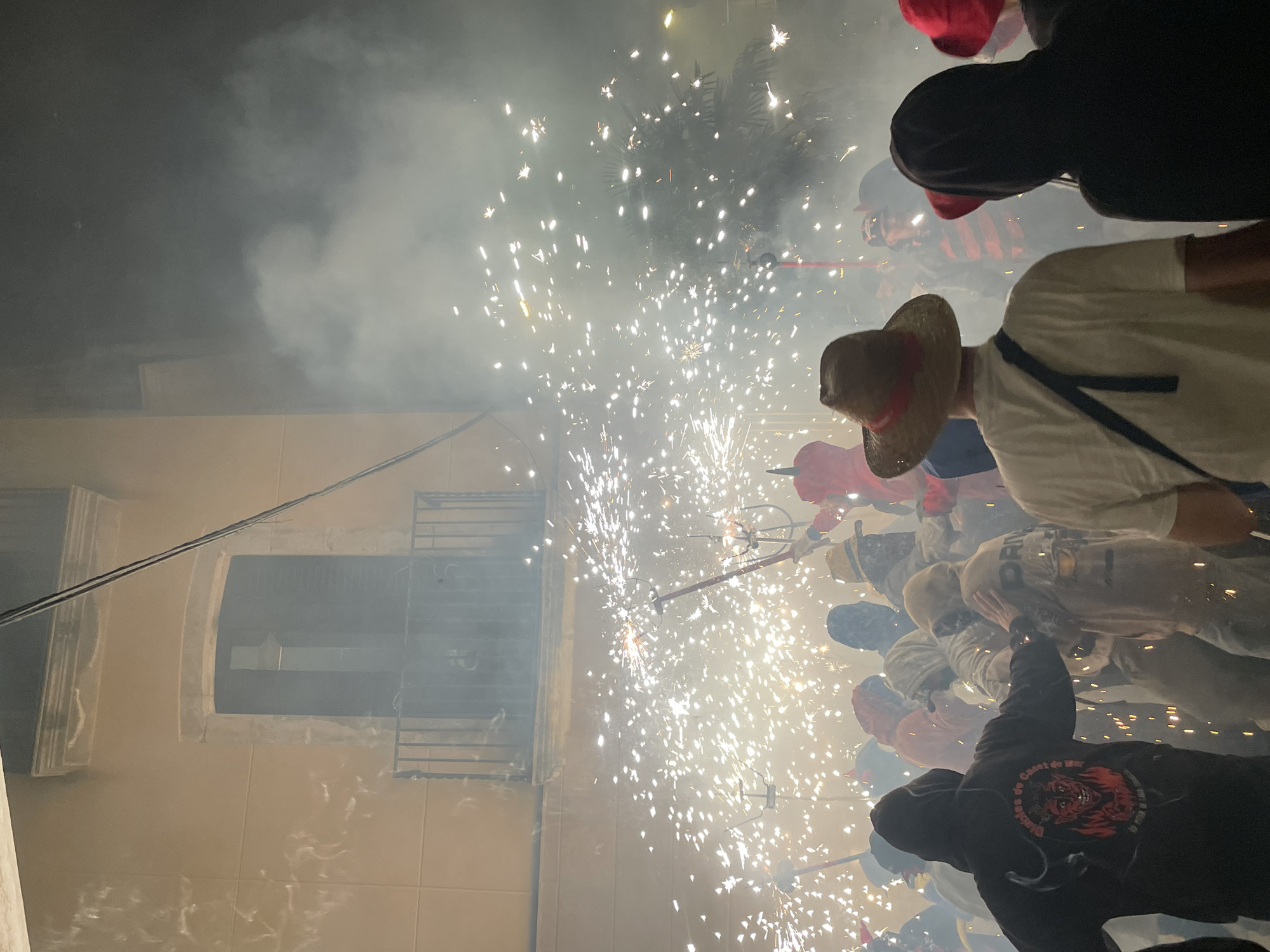 This is a nighttime photograph taken in Canet de Mar, Spain, depicting a lively street event involving fireworks. A dense crowd of people is visible, many wearing hooded sweatshirts and some with hats. The primary action is the display of numerous bright sparks and trails of smoke, indicating that fireworks or pyrotechnic devices are being actively used by participants in the crowd. In the background, a building with a balcony and a window is illuminated by the light of the fireworks, with power lines also visible against the dark sky. The scene is dynamic and energetic, with a strong sense of celebration and community engagement.