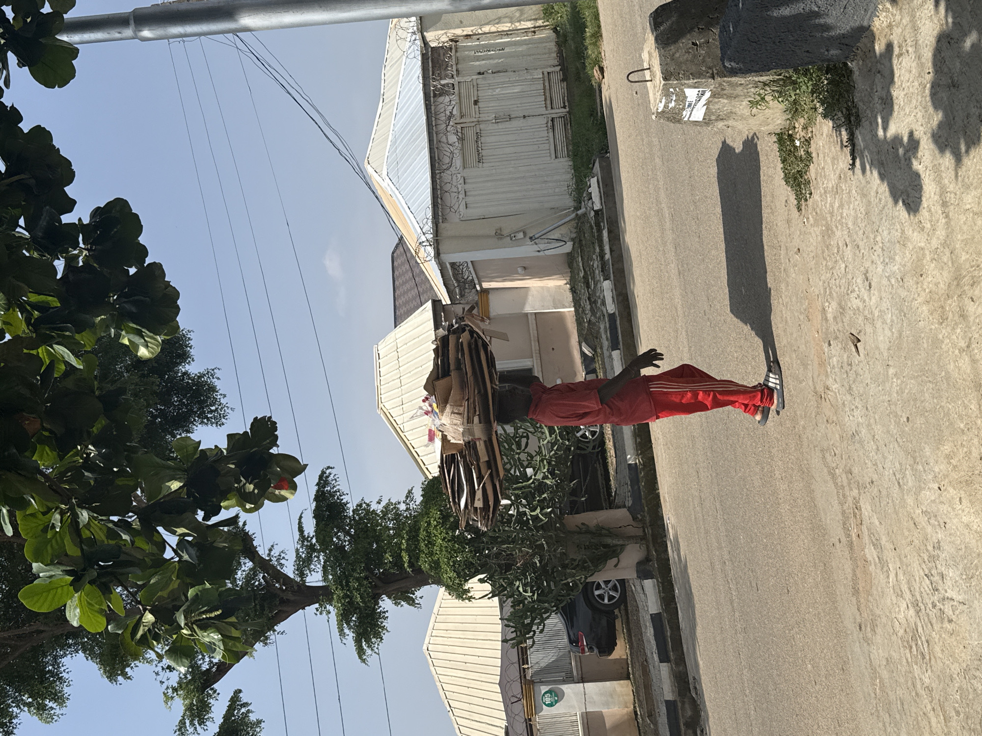 A person wearing red pants with white stripes and black and white athletic shoes is carrying a large bundle of cardboard on their head. They are standing on an asphalt road next to a sidewalk. To the left of the road, there are several buildings with white corrugated metal roofs, some with barbed wire on top. Lush green trees with large leaves are also visible on the left. In the background, there are more buildings and power lines overhead. The sky is clear and blue, suggesting a sunny day. The scene appears to be an urban or semi-urban environment in Abuja, Nigeria, possibly during the daytime. The person's posture and the way they are balancing the load suggest they are in the process of transporting it. There is some graffiti on the buildings, but no clear text is legible in the provided image.