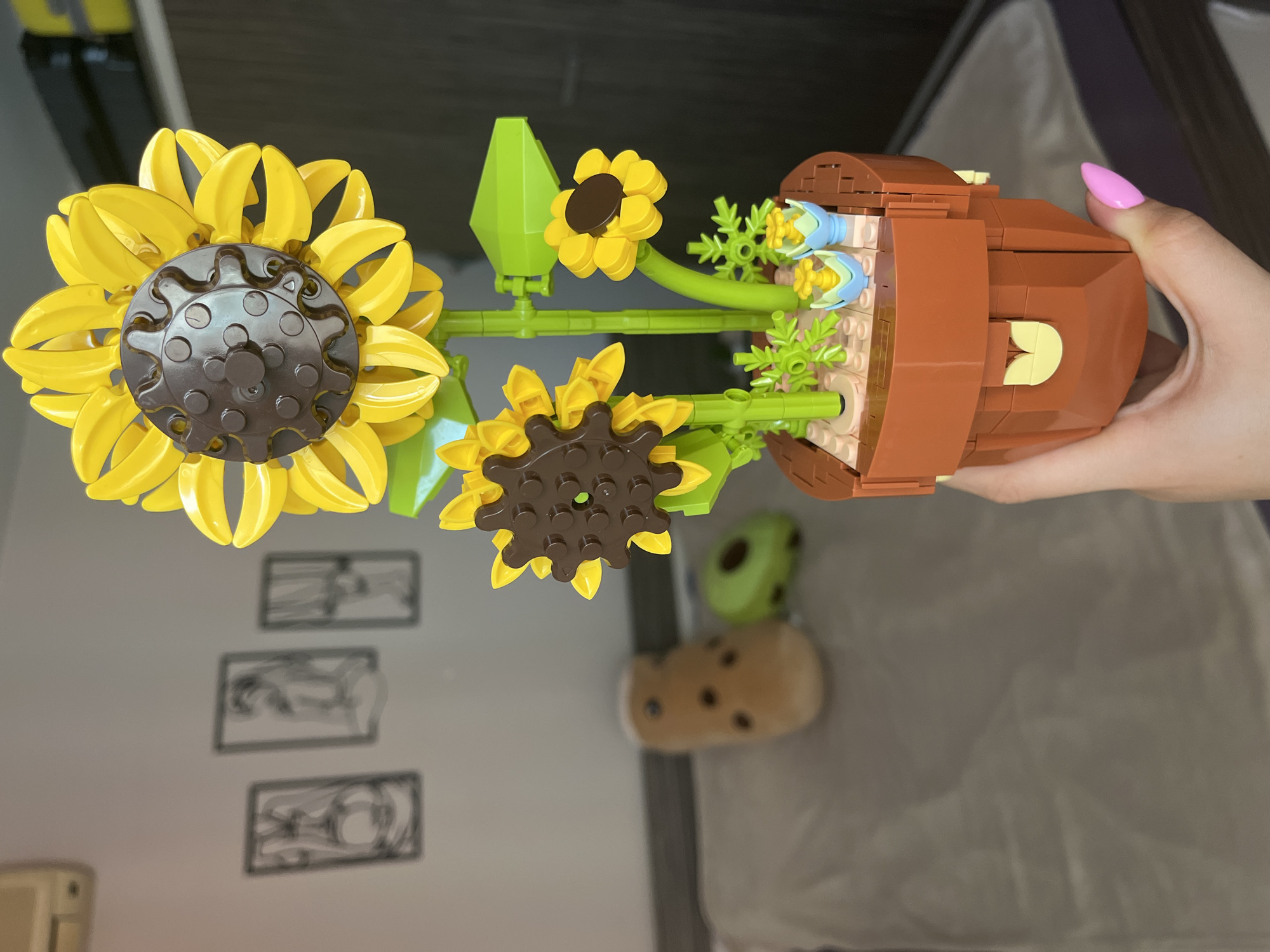 The image shows a LEGO sunflower set in a brown LEGO pot, held by a hand with a pink fingernail. The sunflower has two large yellow blooms with brown centers, a smaller yellow bloom, green stems, and green leaves. The pot is partially filled with light brown and green LEGO pieces. The background includes a wall with three framed, abstract pictures, a tan and green plush toy, part of a bed with a gray and purple blanket, and part of a light-colored cabinet. The image does not provide any information suggesting it was taken in Veliko Tarnovo, Bulgaria.