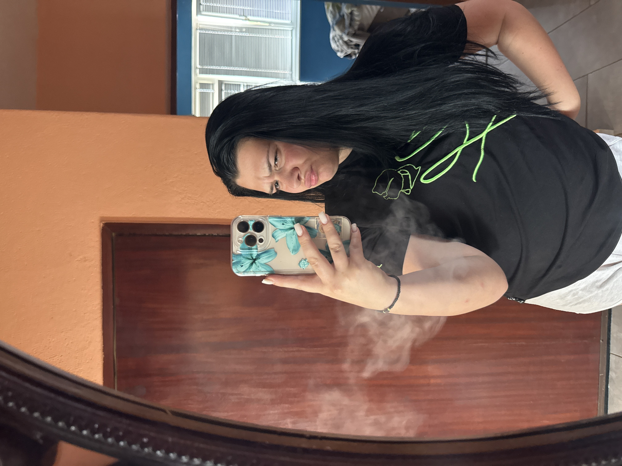 The image depicts a mirror selfie of a young adult female located in the city of Rogosh, Bulgaria. The female has long, dark, straight hair and is wearing a black short-sleeved t-shirt with a neon green outline design on the front, and light grey shorts. She has a pouting or skeptical expression. Her right arm is raised, holding a smartphone with a clear case featuring teal floral patterns. Her nails are painted a light color, possibly white or cream. A black beaded bracelet is visible on her left wrist. A white misty substance is present in the lower-left foreground, partially obscuring her torso. The background reflected in the mirror shows an indoor environment with an orange, textured wall, a dark brown wooden door with a lighter frame, and a section of a blue wall. To the right of the person, a window with white horizontal blinds is visible, along with a glimpse of what appears to be furniture with a patterned covering. The floor appears to be light-colored tile.