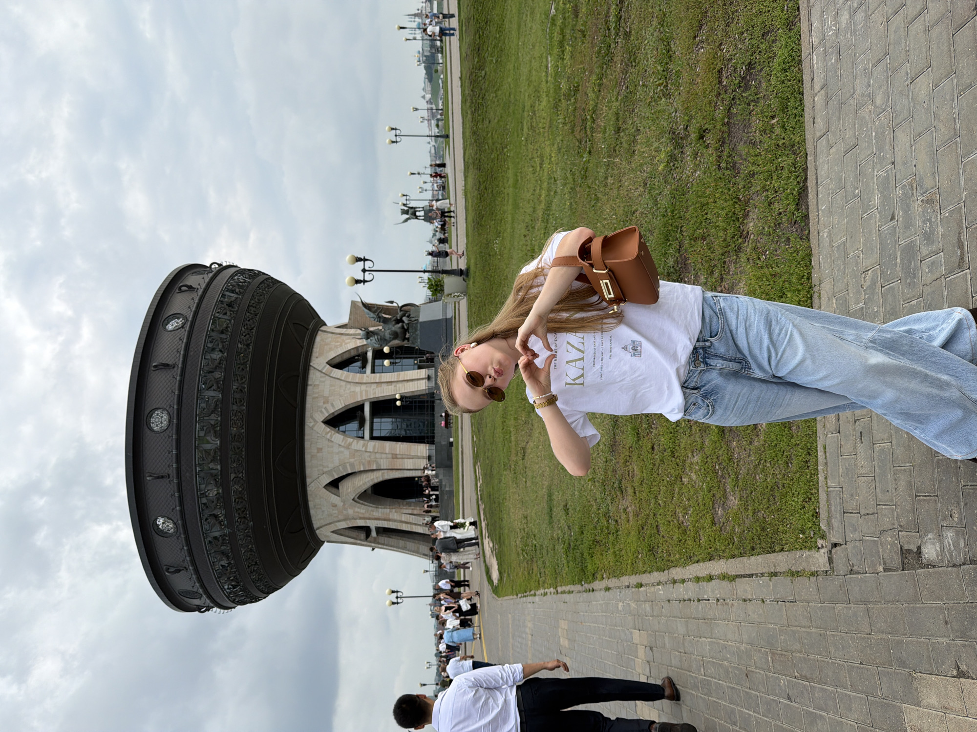 In Kazan, Russia, a young woman with blonde hair and sunglasses stands in the foreground, making a heart shape with her hands. She wears a white t-shirt with text and light blue wide-leg jeans, and carries a brown handbag. Behind her, a man in a white shirt and dark pants walks away from the camera on a paved path. In the background, a large, ornate cauldron-shaped structure with arched openings and decorative metalwork stands prominently against a cloudy sky. The area around the structure is a grassy park with lamp posts and several people milling about, some appearing to be attending an event, possibly a wedding, near the entrance of the building. Further in the distance, the Kazan Kremlin's spires and rooftops are visible.