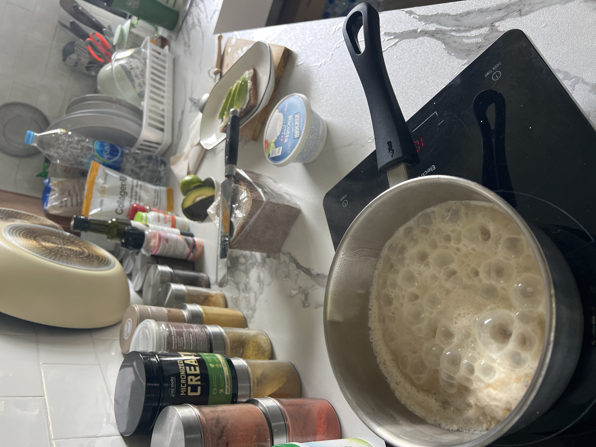 A close-up shot taken at an angle in a kitchen setting. The scene is tightly packed, with various items filling the space.
On the left side of the frame, a rack is visible, holding dishes, kitchen tools like scissors, and other utensils. A variety of containers and bottles, including those with labels such as "Collagen" and "micronized creatine", are stacked. A frying pan is also present.
Moving towards the center, there is a plate with what appears to be avocado on toast, along with a container labeled "Ricotta". Next to it, a loaf of bread is partially wrapped in plastic.
On the right side, a stainless steel saucepan sits on a black induction cooktop. The contents of the saucepan are bubbly, suggesting something is being cooked. The cooktop has power and timer controls visible.
The background features a mottled white surface, giving the impression of a marble countertop. The overall atmosphere is that of a cluttered but functional kitchen space.
The location in the city of บ้านบางรักษ์, Thailand is not determinable from the image.