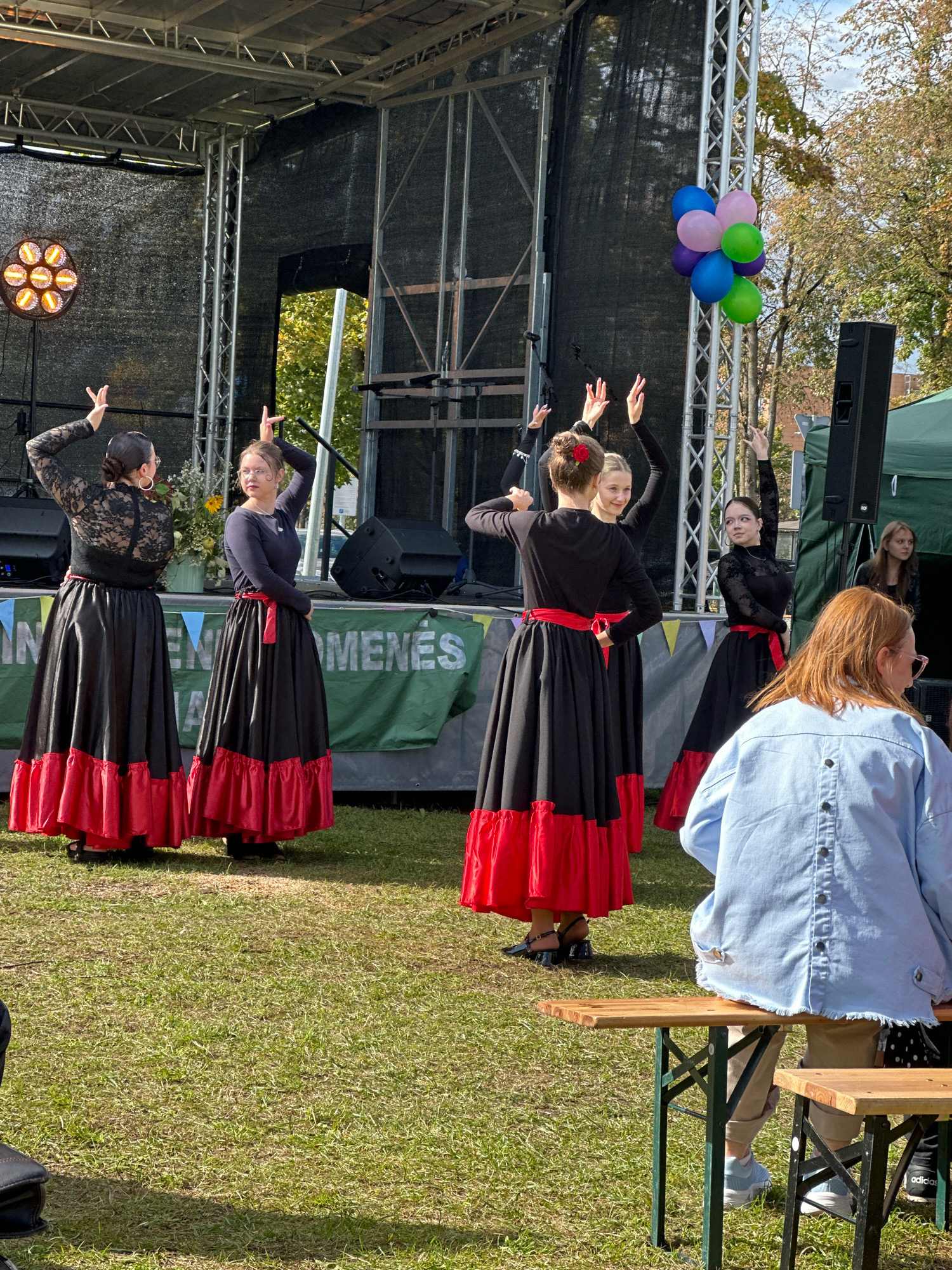 The image depicts an outdoor performance on a stage set up on a grassy area. The stage has a black backdrop and is adorned with lighting fixtures. In the foreground, five young women are performing in traditional black and red folk dresses with ruffled skirts. They are facing away from the camera, their arms raised in a synchronized dance. Behind them, a cluster of colorful balloons, including blue, purple, pink, and green, are tied to a metal structure. Spectators are seated at picnic tables in the foreground, with one woman in a light blue denim jacket facing away from the camera, looking towards the stage. The background shows trees with autumn foliage and a clear sky. The presence of the "Vilnius - Lietuvos sostinė" banner on the stage suggests the performance is taking place in Vilnius, Lithuania.