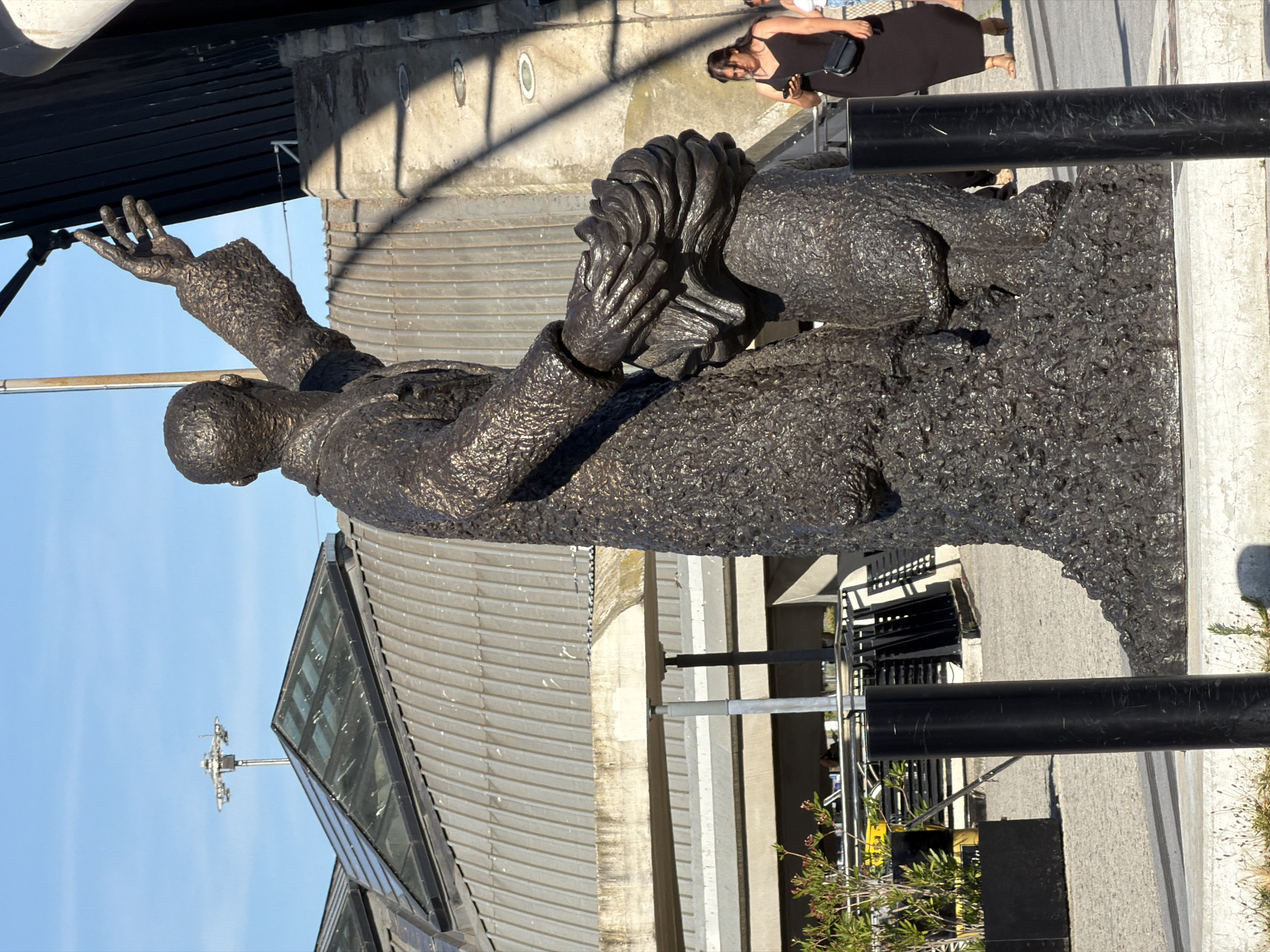 This media file is a portrait-oriented photograph of a dark, textured bronze statue of a person from the chest up, viewed from a slightly elevated, right-facing angle. The statue appears to be of a man wearing a suit and tie, with his head tilted back and his left arm raised with an open palm facing upwards. His right arm is bent, with his hand resting on his chest or head. The statue has a rough, almost granular surface texture.
In the background, to the upper right of the statue, a woman in a dark dress is partially visible, holding a small black purse. She is looking down and to her left. The background also includes architectural elements such as concrete structures, metal railings, and a sloped, grey roof with a glass section. A black cylindrical pole runs vertically in the foreground, partially obscuring the statue.
The scene is outdoors, under a clear blue sky, suggesting it is daytime with sunny weather. The lighting casts shadows on the concrete structures, indicating the sun is at an angle. The overall mood is one of grandeur and perhaps contemplation, conveyed by the statue's pose. The image is taken in Colombier-Saugnieu, France. There is no visible text in the image.