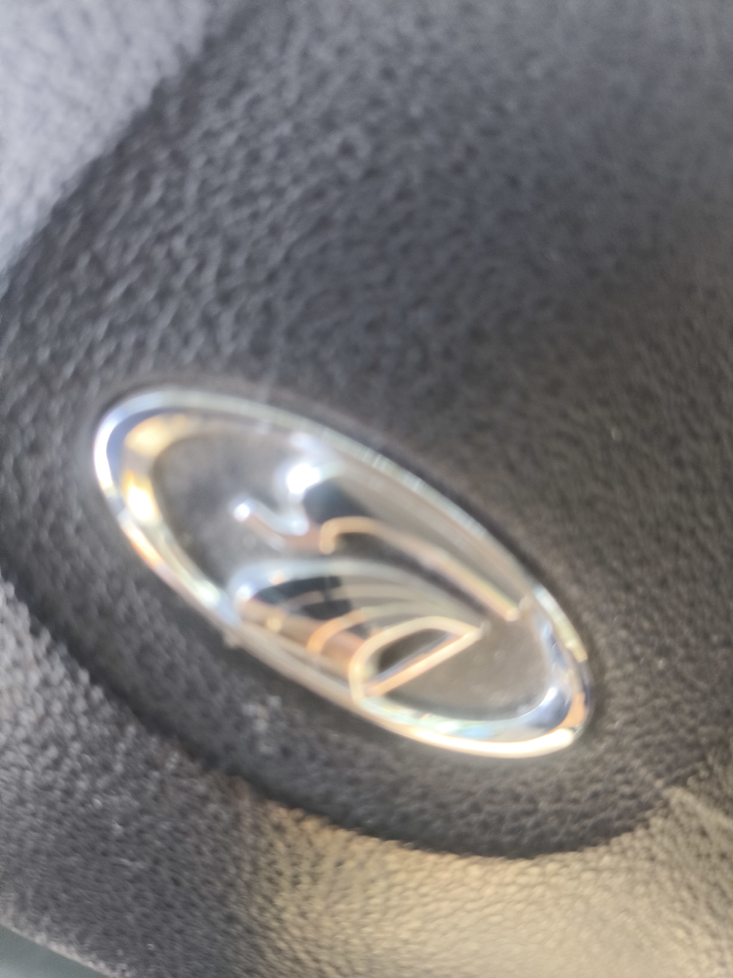 The image shows a close-up, slightly out-of-focus view of a chrome emblem with a reflective surface on a textured, dark grey material, likely leather, consistent with a car's steering wheel. The emblem appears to be a stylized shape with curved lines, possibly a logo. The lighting creates bright reflections and highlights on the emblem. There are no people or any identifiable city landmarks present. The setting is domestic, within a vehicle.