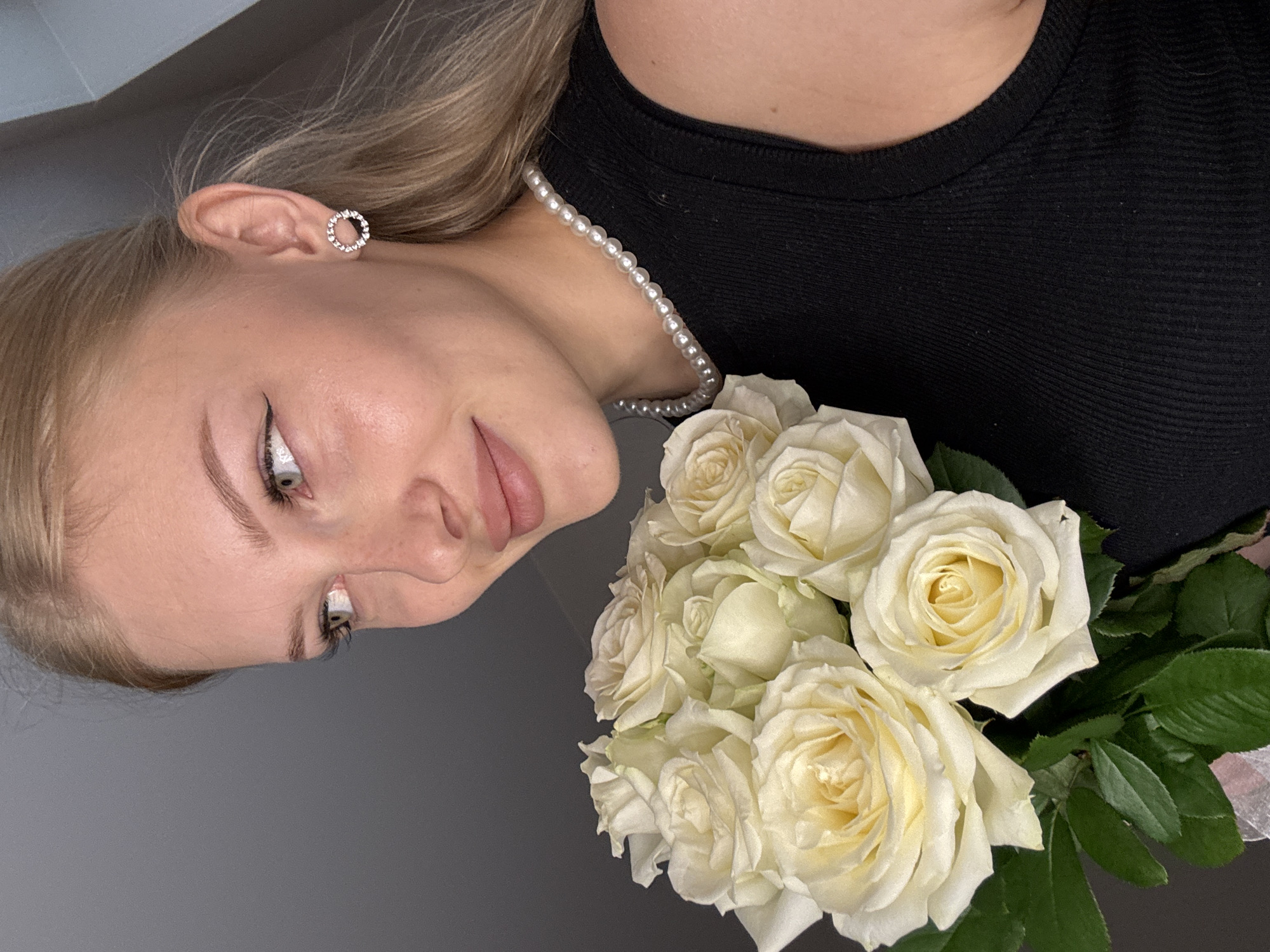 A young woman with blonde hair pulled back, a black ribbed tank top, and a pearl necklace is holding a bouquet of white roses. She is wearing circular earrings with rhinestones and has winged eyeliner. The background is a plain grey wall. The image appears to be a selfie taken indoors, with the woman looking off to the side. There is no indication of the city of Perm, Russia, in the image.