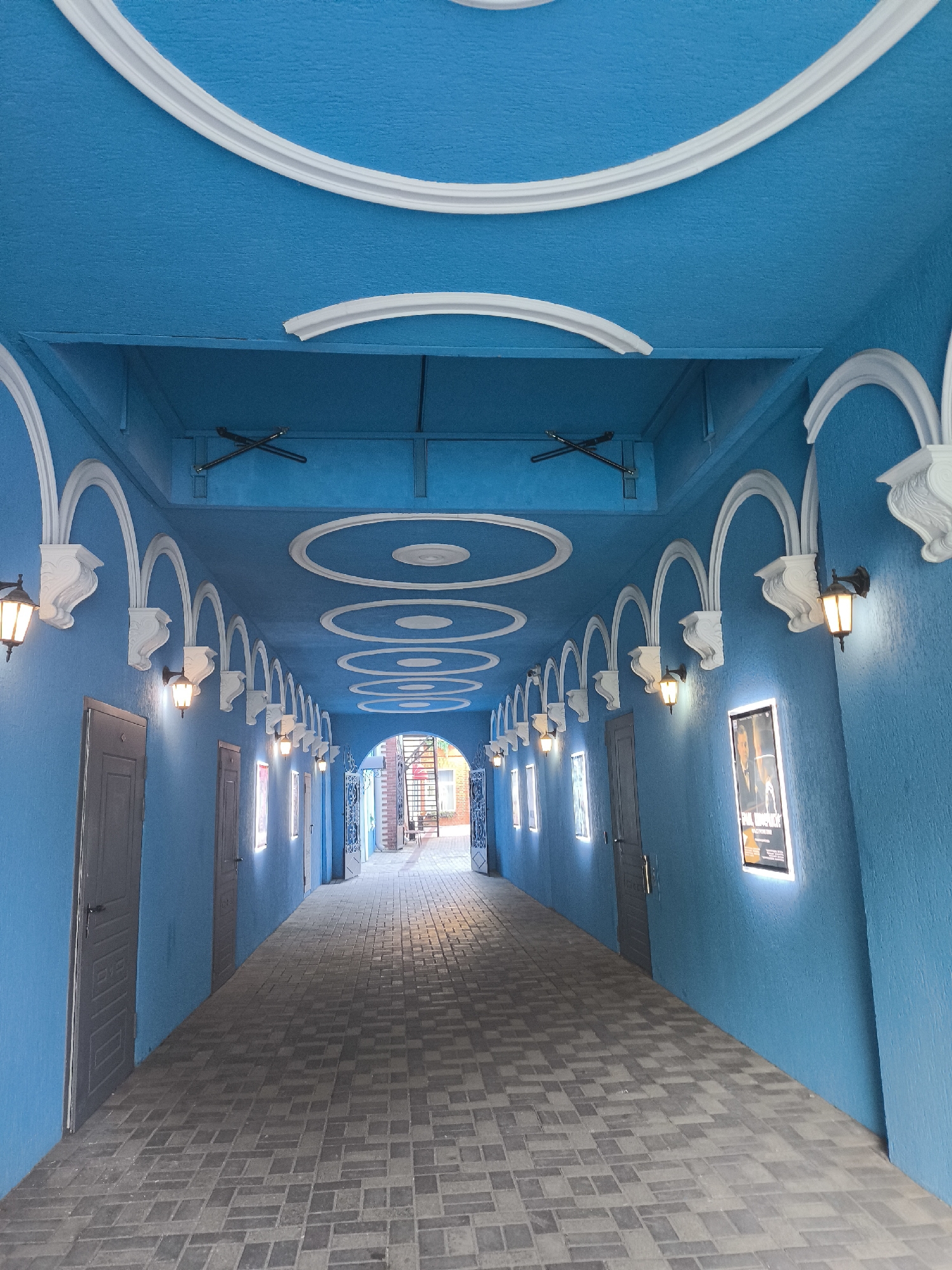 The image depicts a bright blue covered walkway or corridor with decorative white molding on the ceiling, which features a repeating pattern of concentric circles. The walls are also painted blue and are adorned with arched white decorative elements extending from the ceiling. Along the left wall, there are several gray doors and illuminated rectangular frames, possibly for posters or art. Along the right wall, there are similar gray doors and illuminated frames, including one with a poster featuring two men. Several vintage-style wall lamps with warm light are mounted on the arched decorative elements, casting a glow. The walkway is paved with gray rectangular bricks laid in a pattern. The corridor recedes into the distance, leading to an open area with more buildings visible. There are no people present in the image. The overall impression is of a creatively decorated, perhaps theatrical or entertainment-themed, public space. This location is identified as a corridor in the city of Perm, Russia.